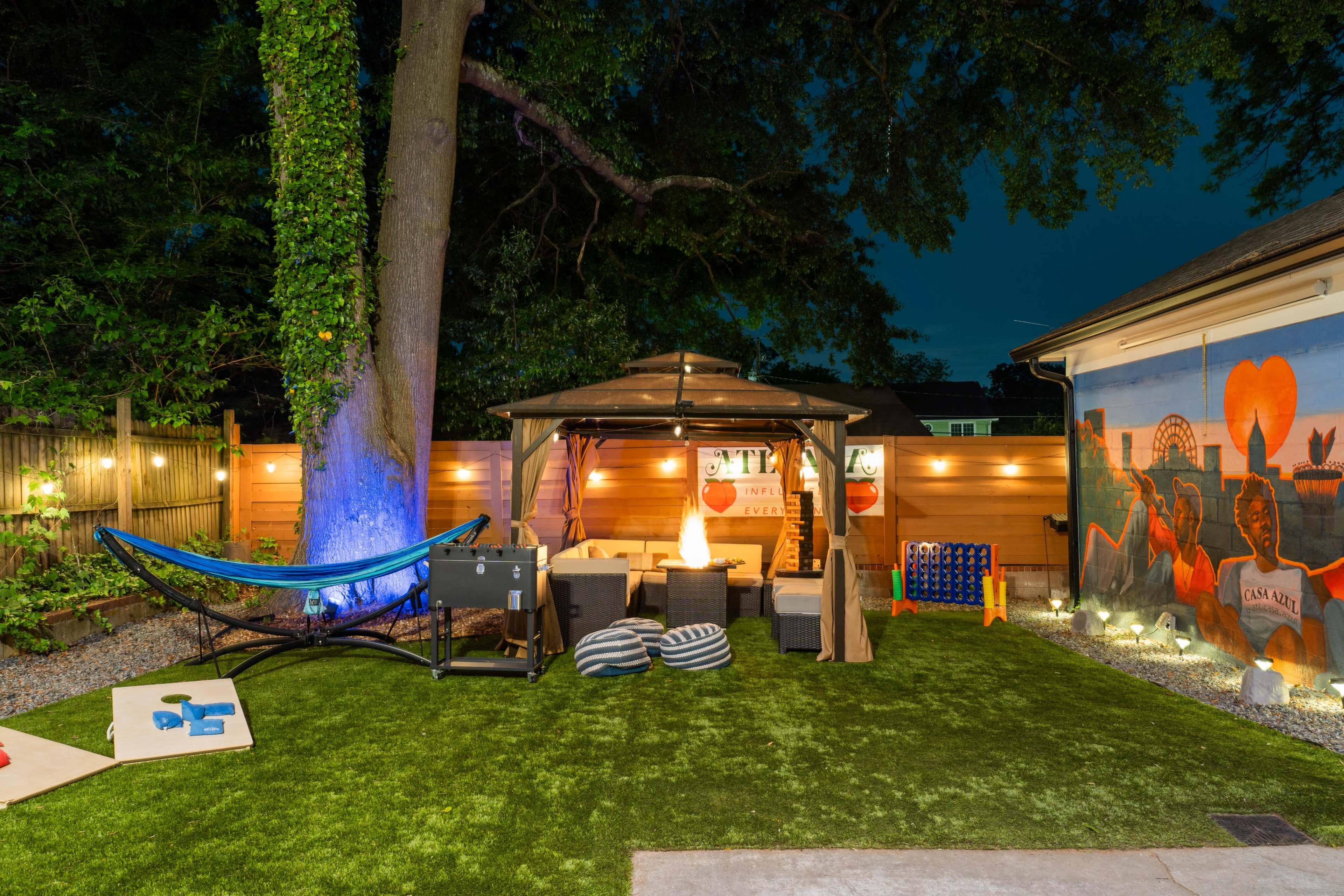 The image shows a backyard scene featuring a seating area under a gazebo, a hammock, and a mural on a wall, all illuminated by string lights at dusk.