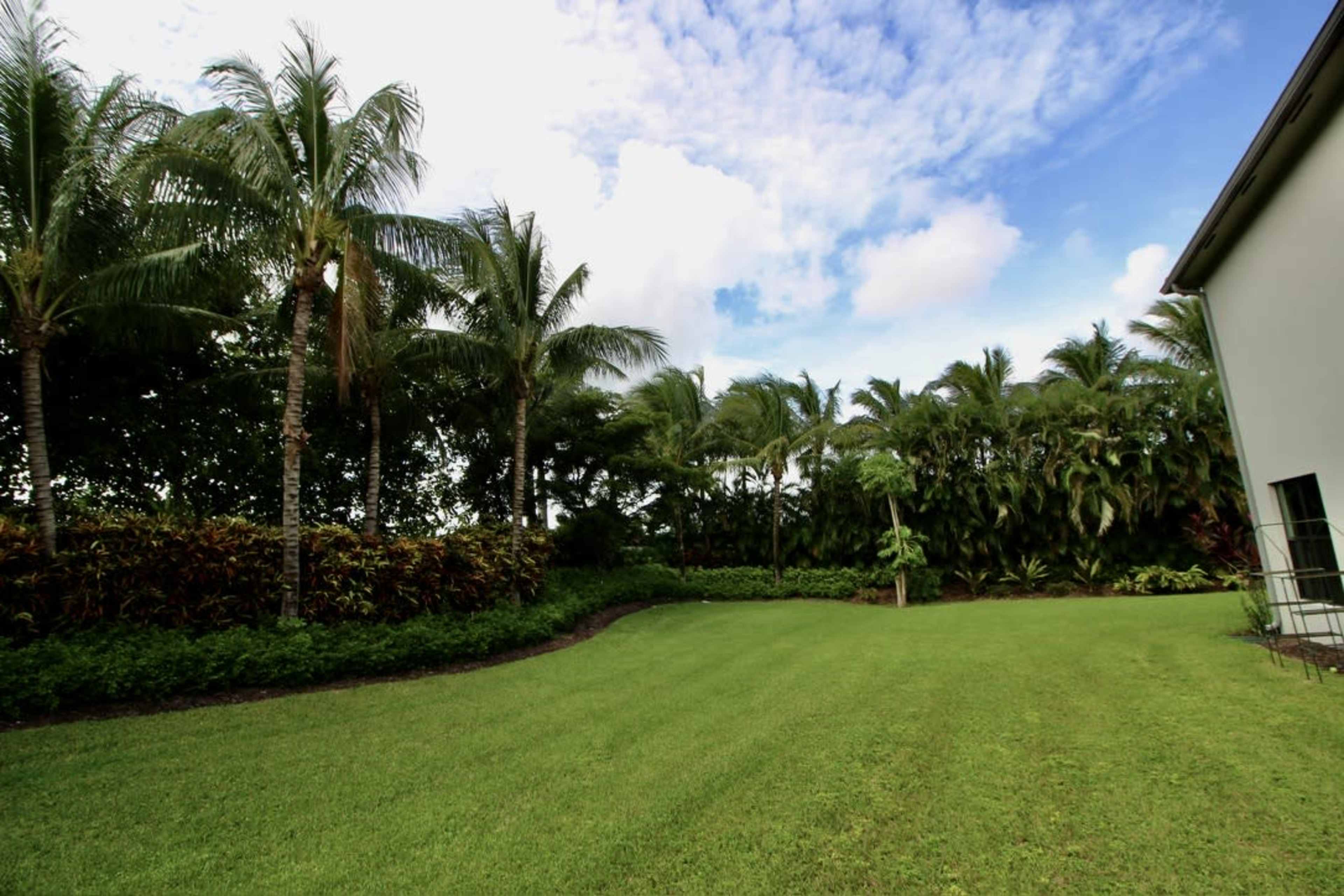 A well-maintained lawn is bordered by tropical trees and shrubs under a partly cloudy sky.