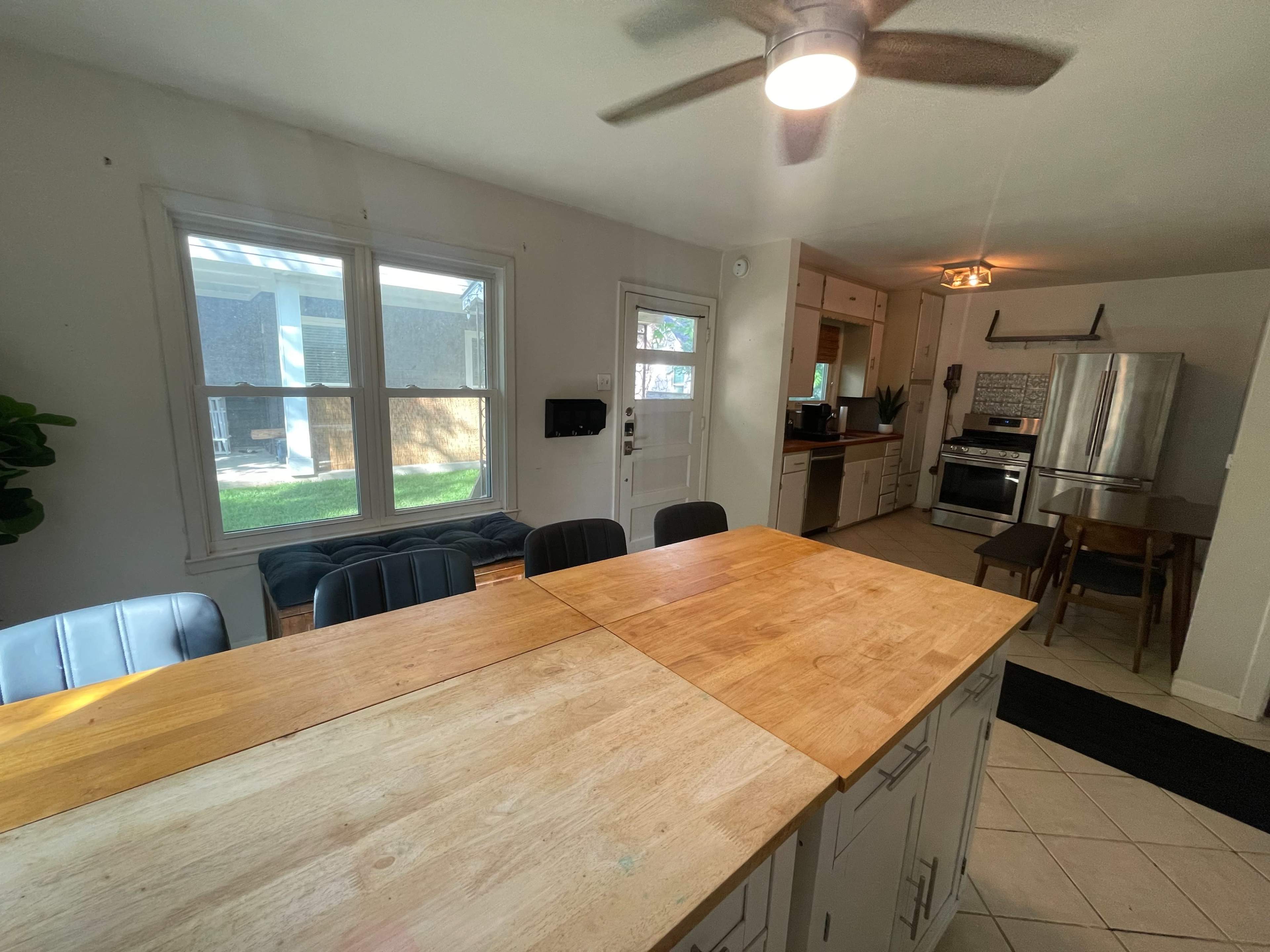 The image shows a kitchen and dining area with a wooden table, chairs, and modern appliances, illuminated by natural light from a window.