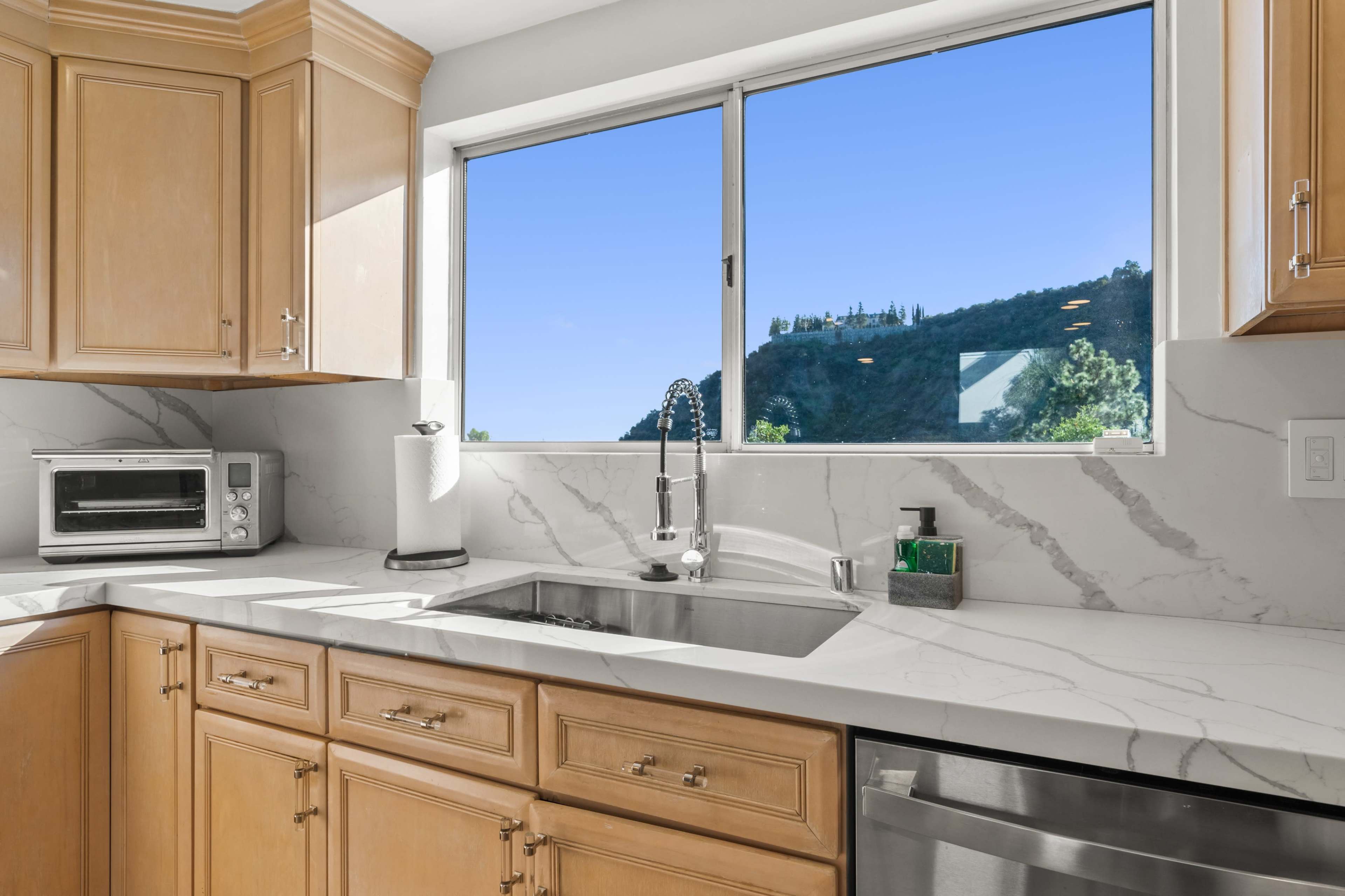 A bright kitchen with light wooden cabinets, a marble countertop, and a window overlooking a green hillside.