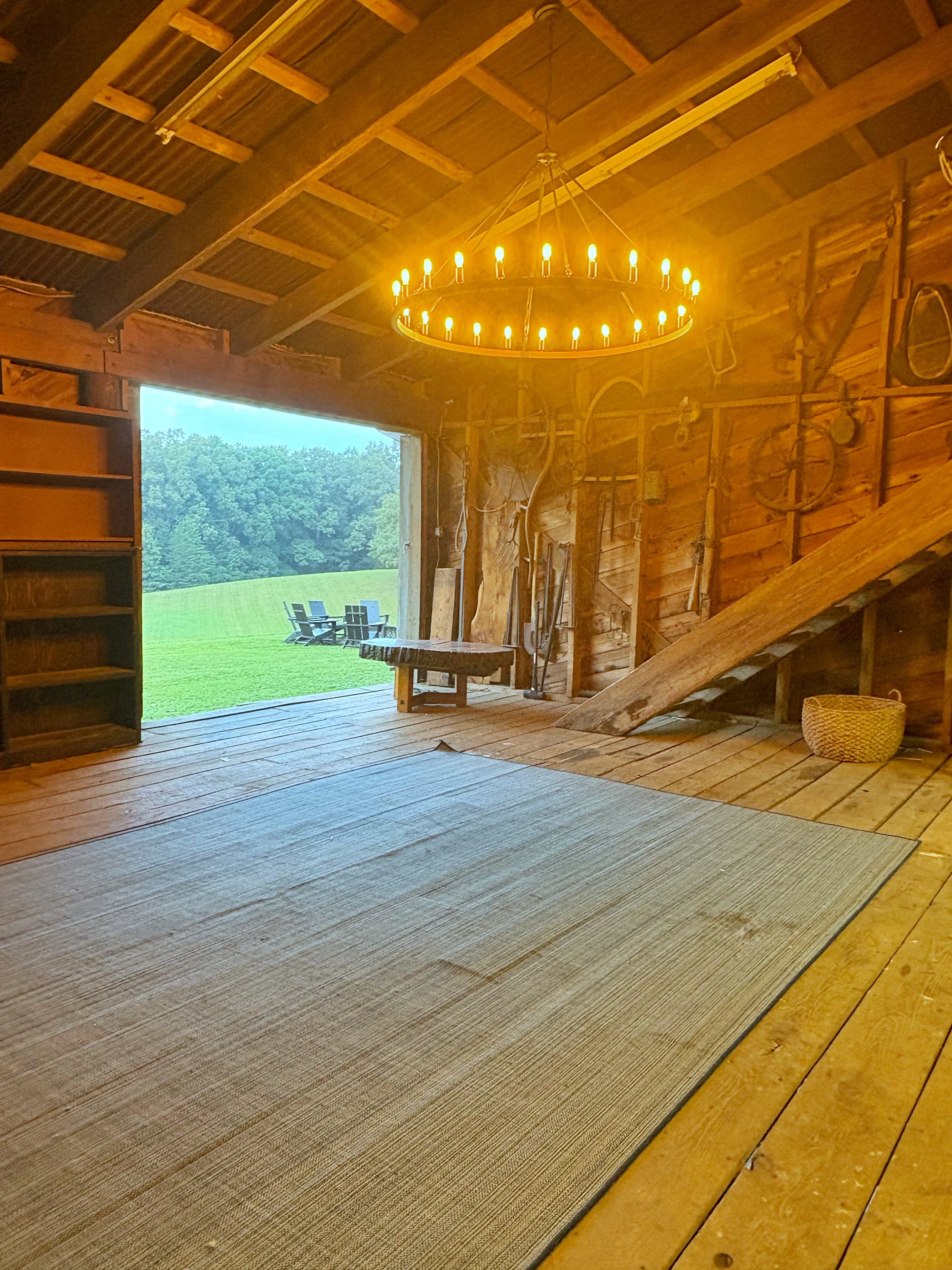 The interior of a rustic barn features wooden beams, a large chandelier, and an open entrance leading to a grassy landscape with outdoor seating.