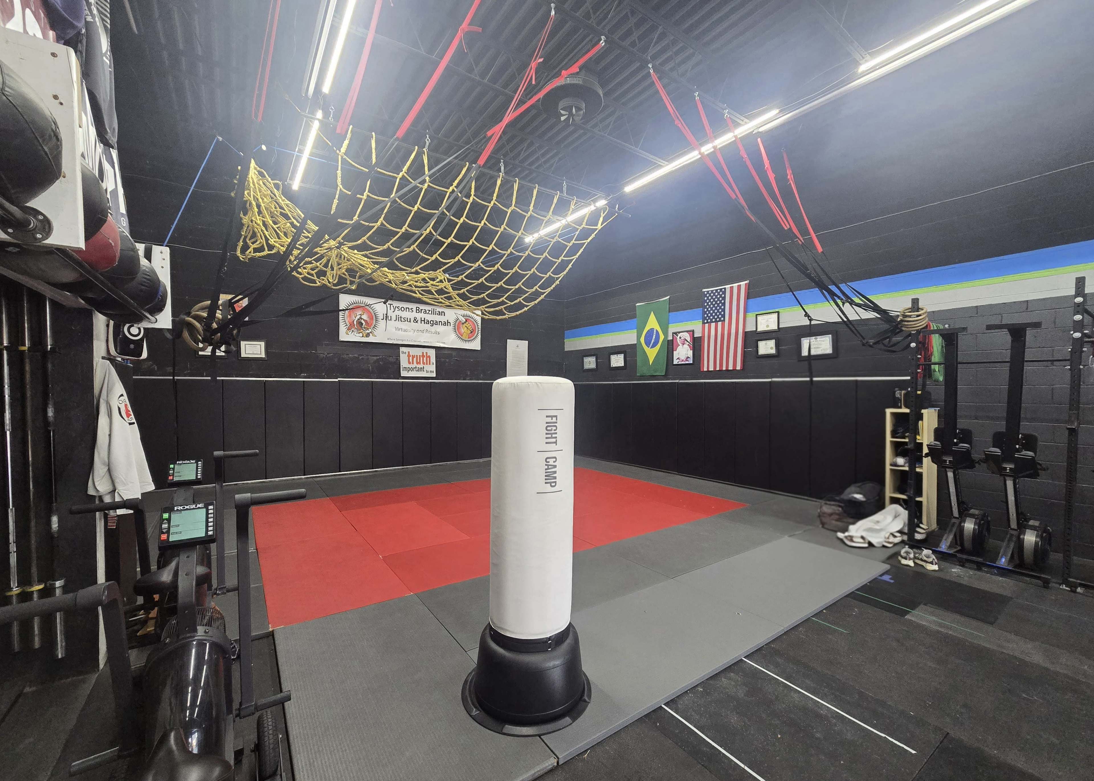 The image shows a training facility with a red mat area, a punching bag, various gym equipment, and several flags displayed on the walls.