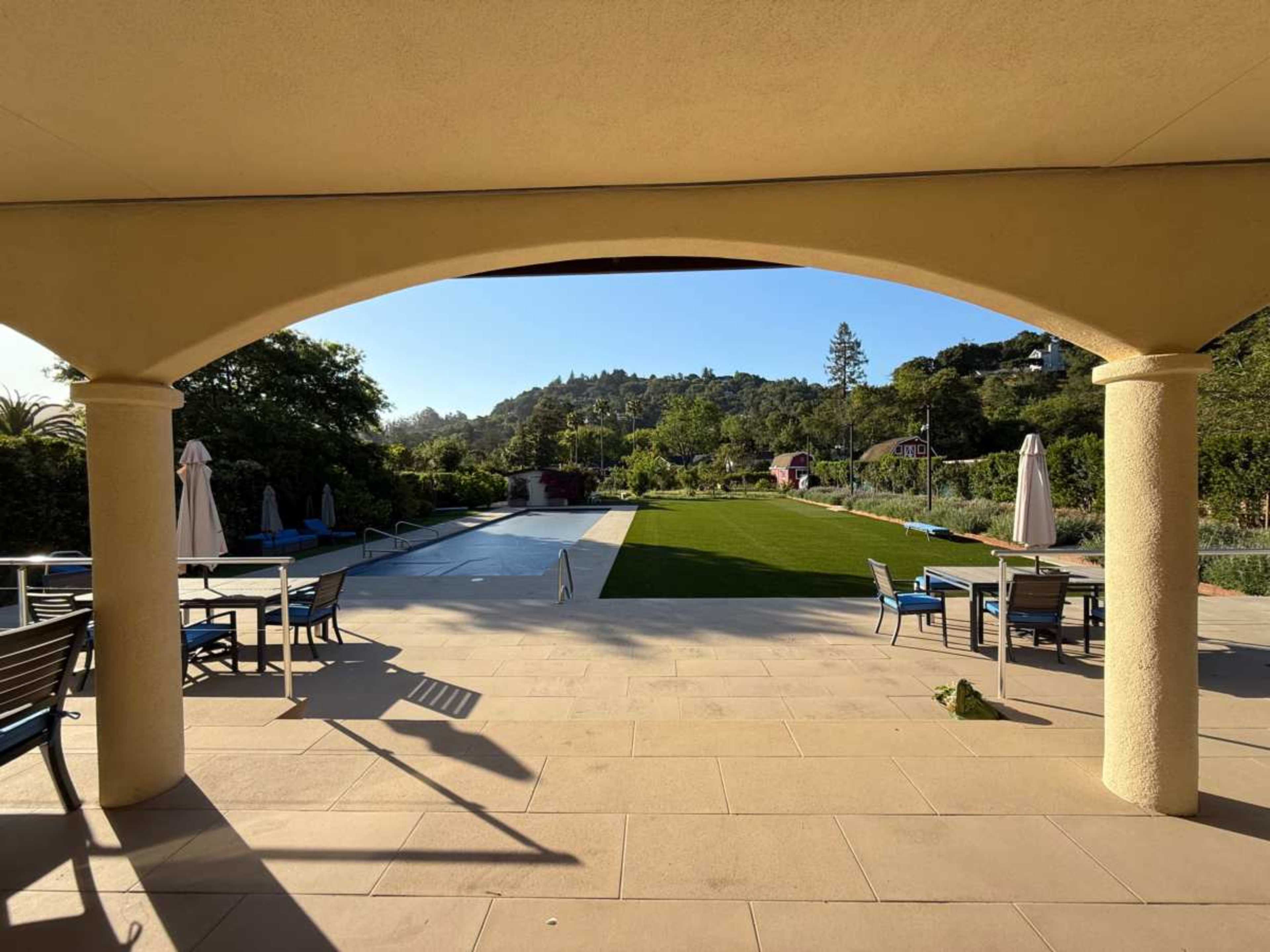 The view from a shaded patio shows a pool area bordered by a green lawn and trees in the background.