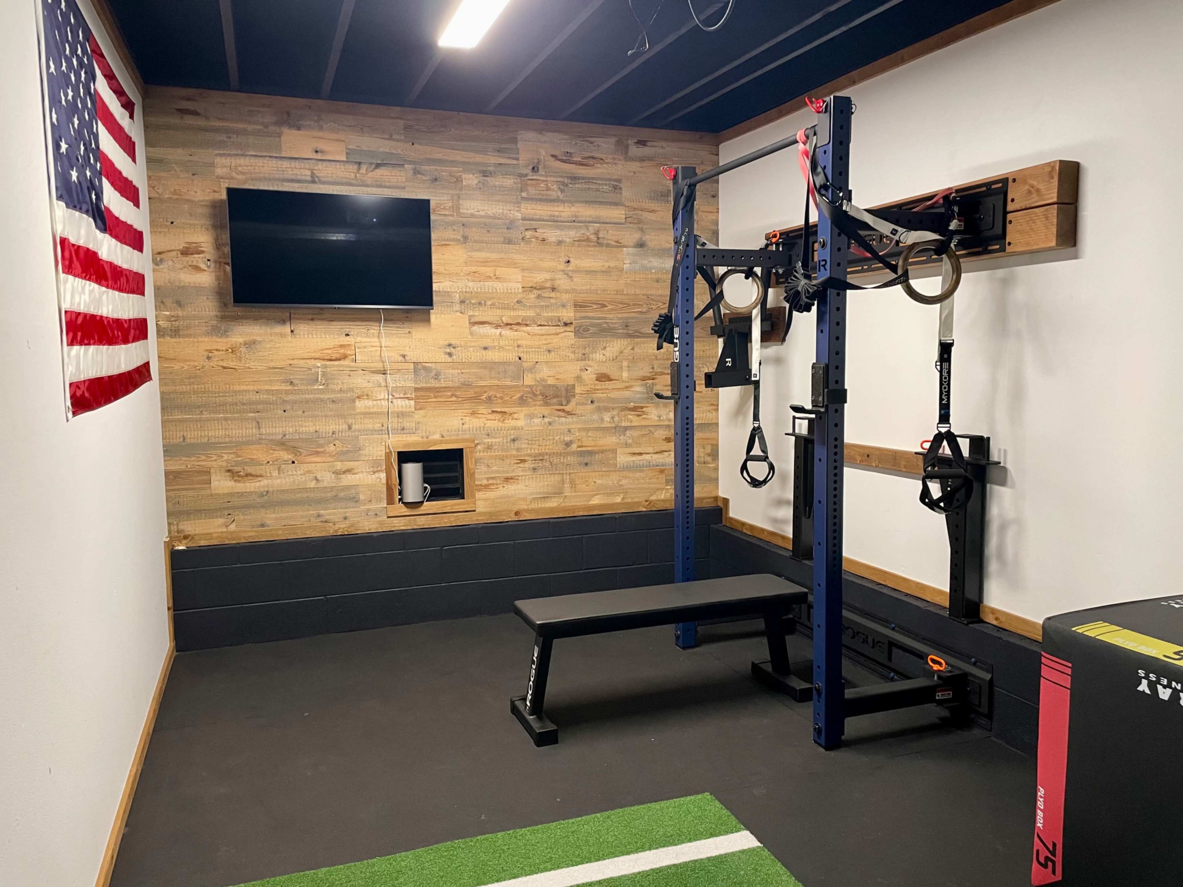 A home gym featuring a power rack with pull-up bars, suspension straps, a bench, a wall-mounted TV, and an American flag on the wall.