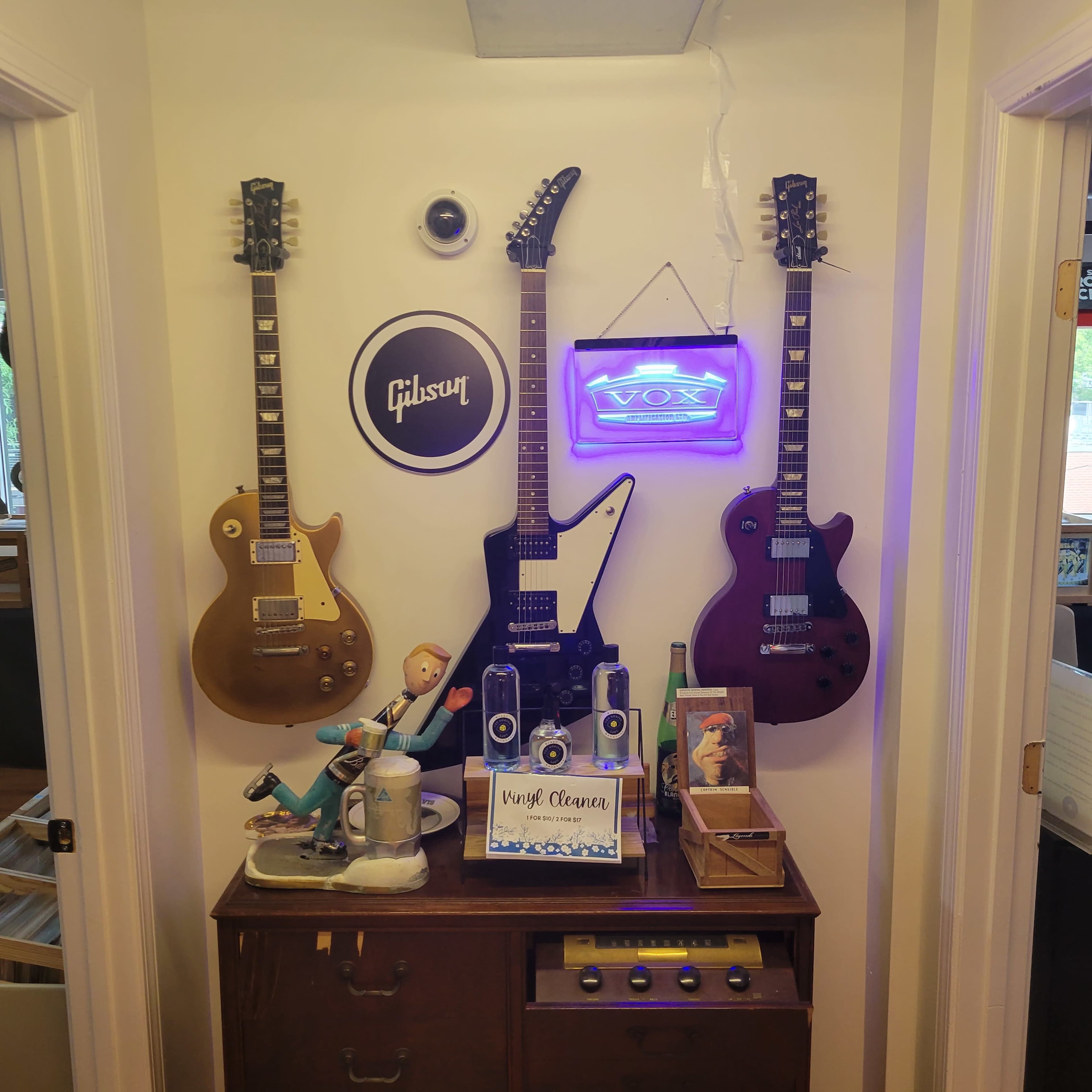 A wall display with three guitars hanging, a neon sign that reads "VOX," and a vintage-style table with a small figurine and speakers.