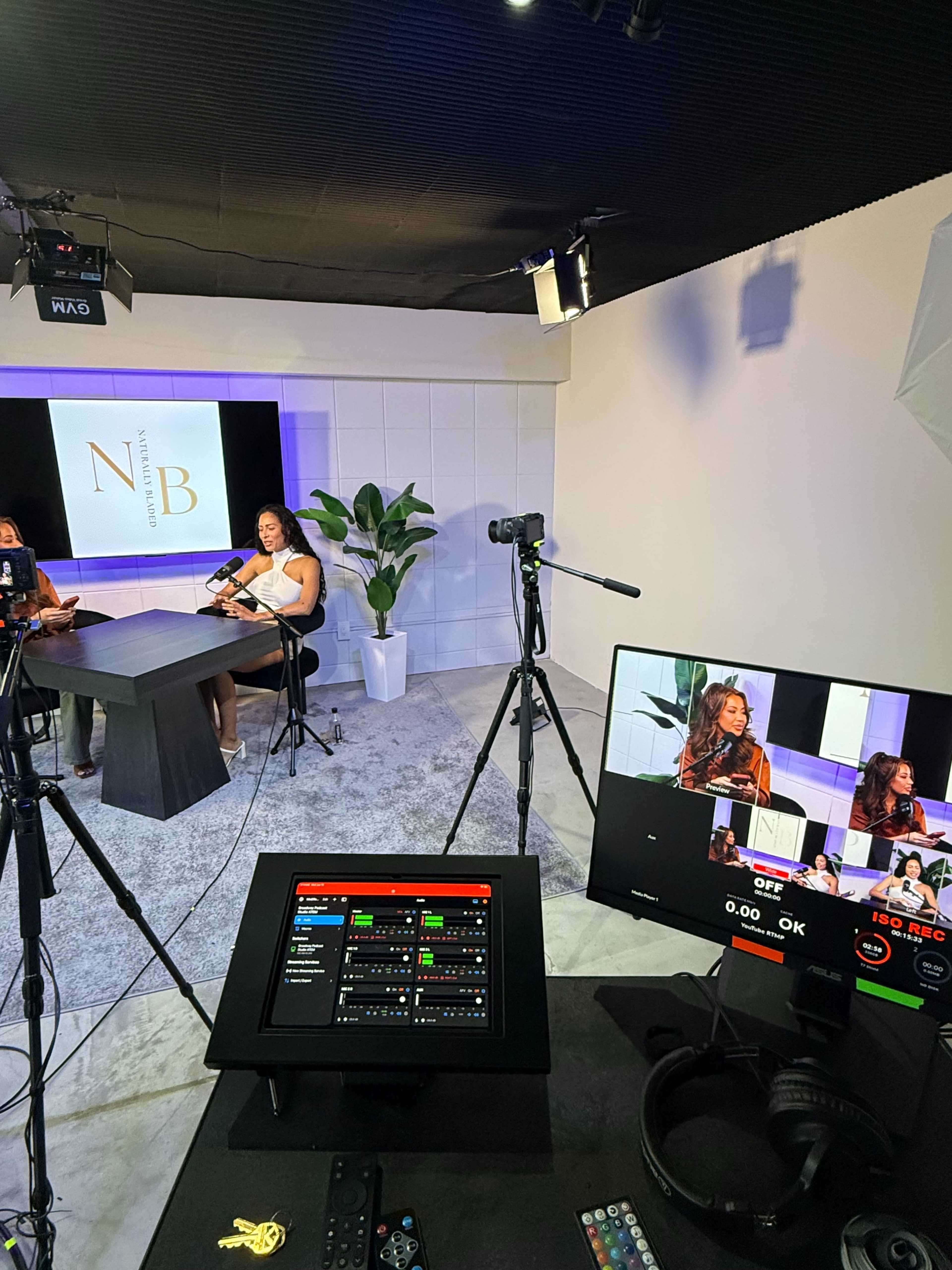 A media studio setup features two women sitting at a table with microphones, surrounded by cameras and lighting equipment, alongside a large monitor displaying their images.
