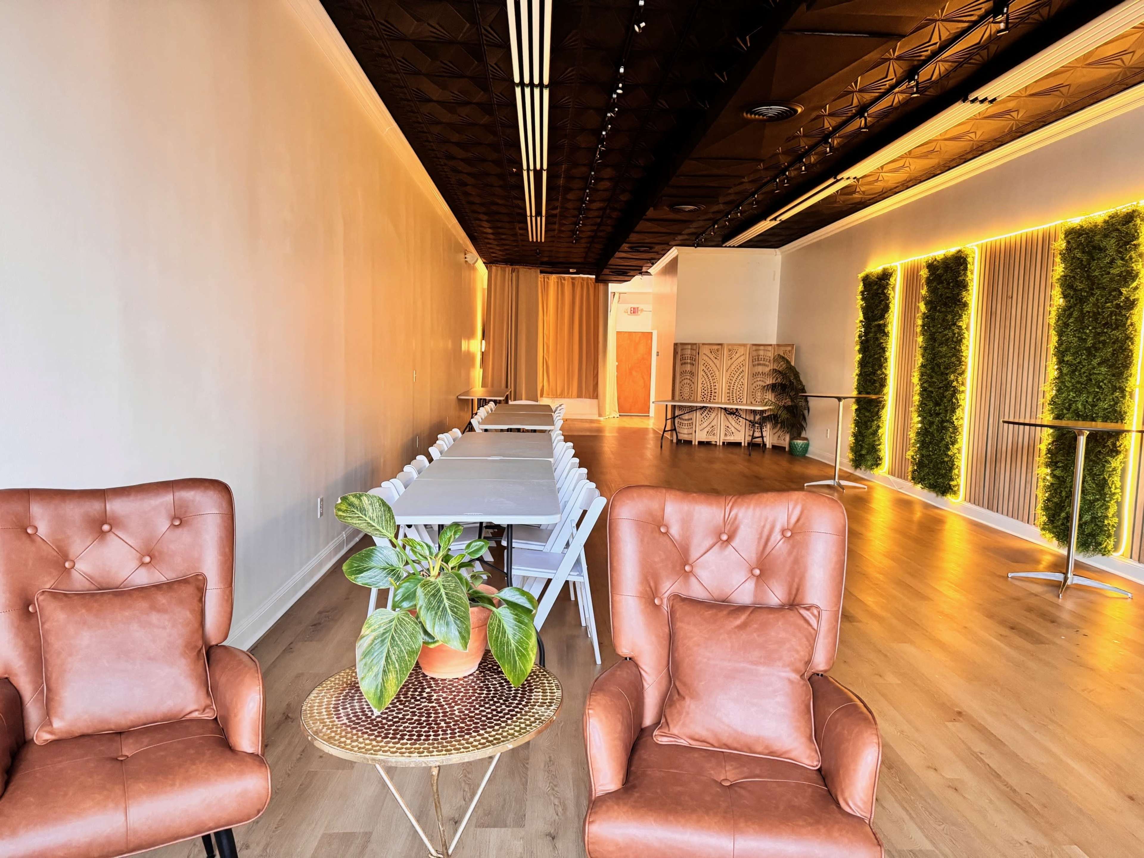The image shows a spacious interior with brown armchairs, a row of tables and chairs, and wall-mounted greenery lighting.