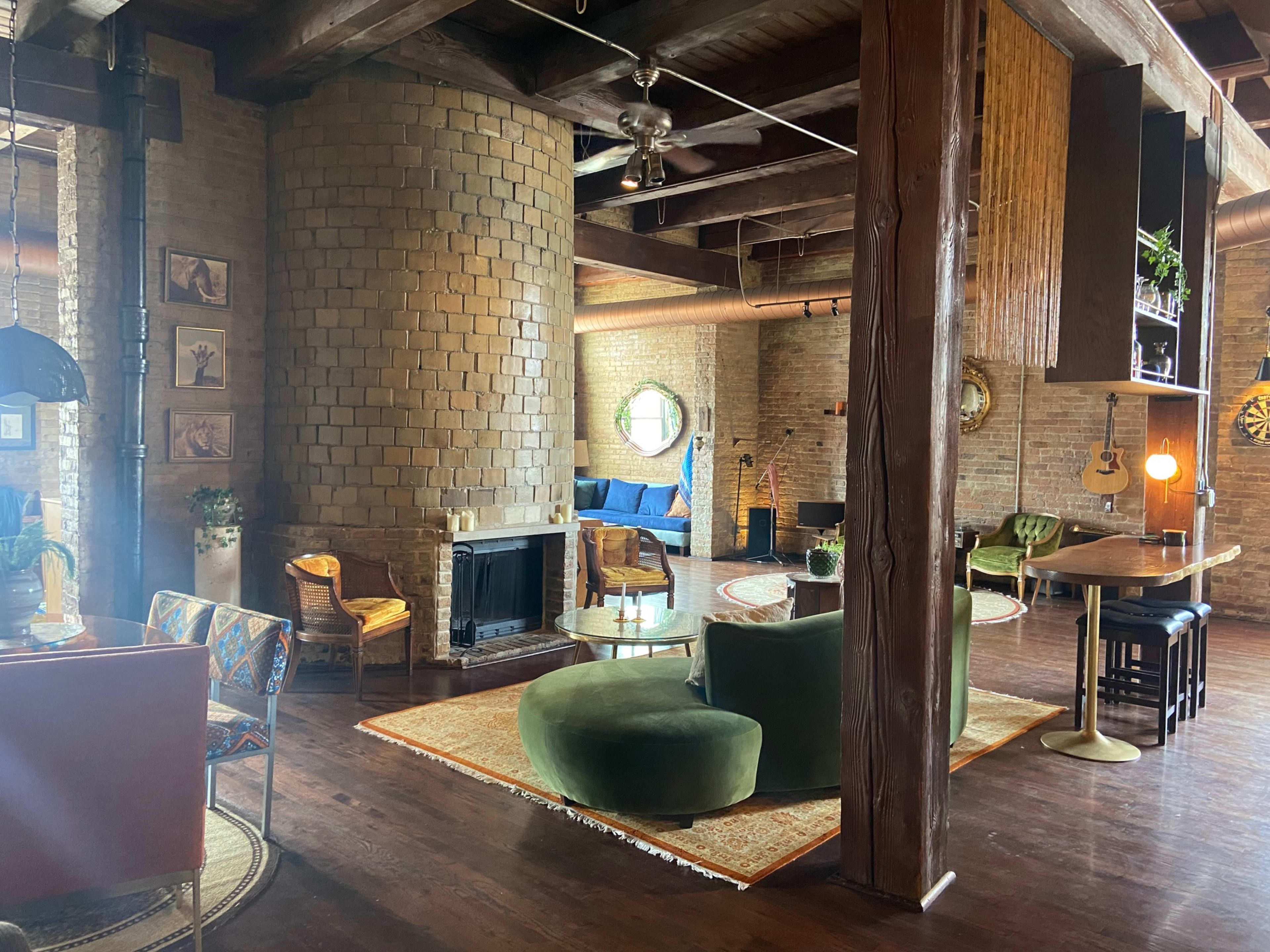 The interior features a spacious room with exposed brick walls, wooden beams, and a circular fireplace, surrounded by various seating areas and decorative elements.