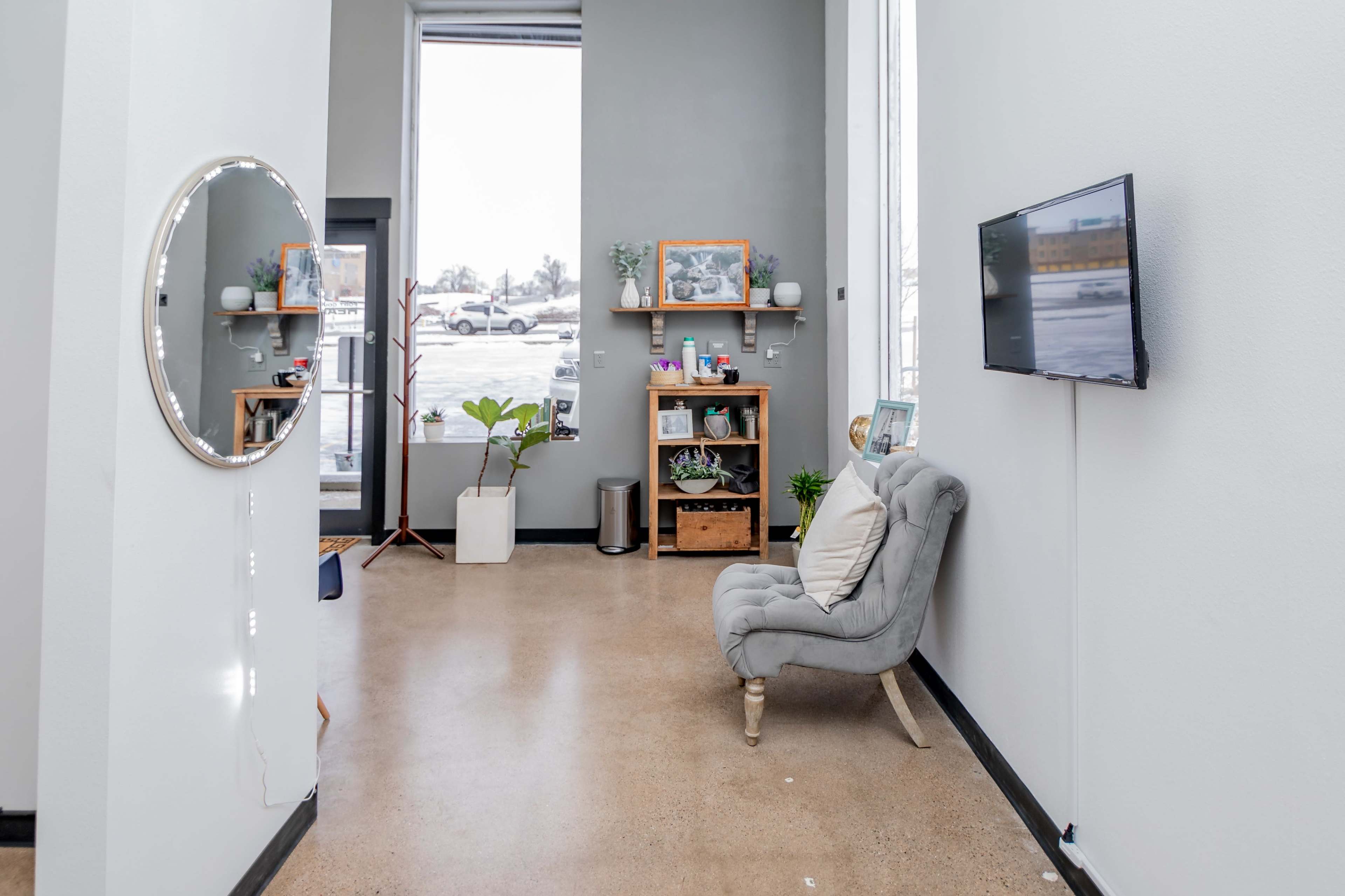 The image shows a modern interior space with a gray armchair, a small shelf displaying various items, a television mounted on the wall, and large windows letting in natural light.