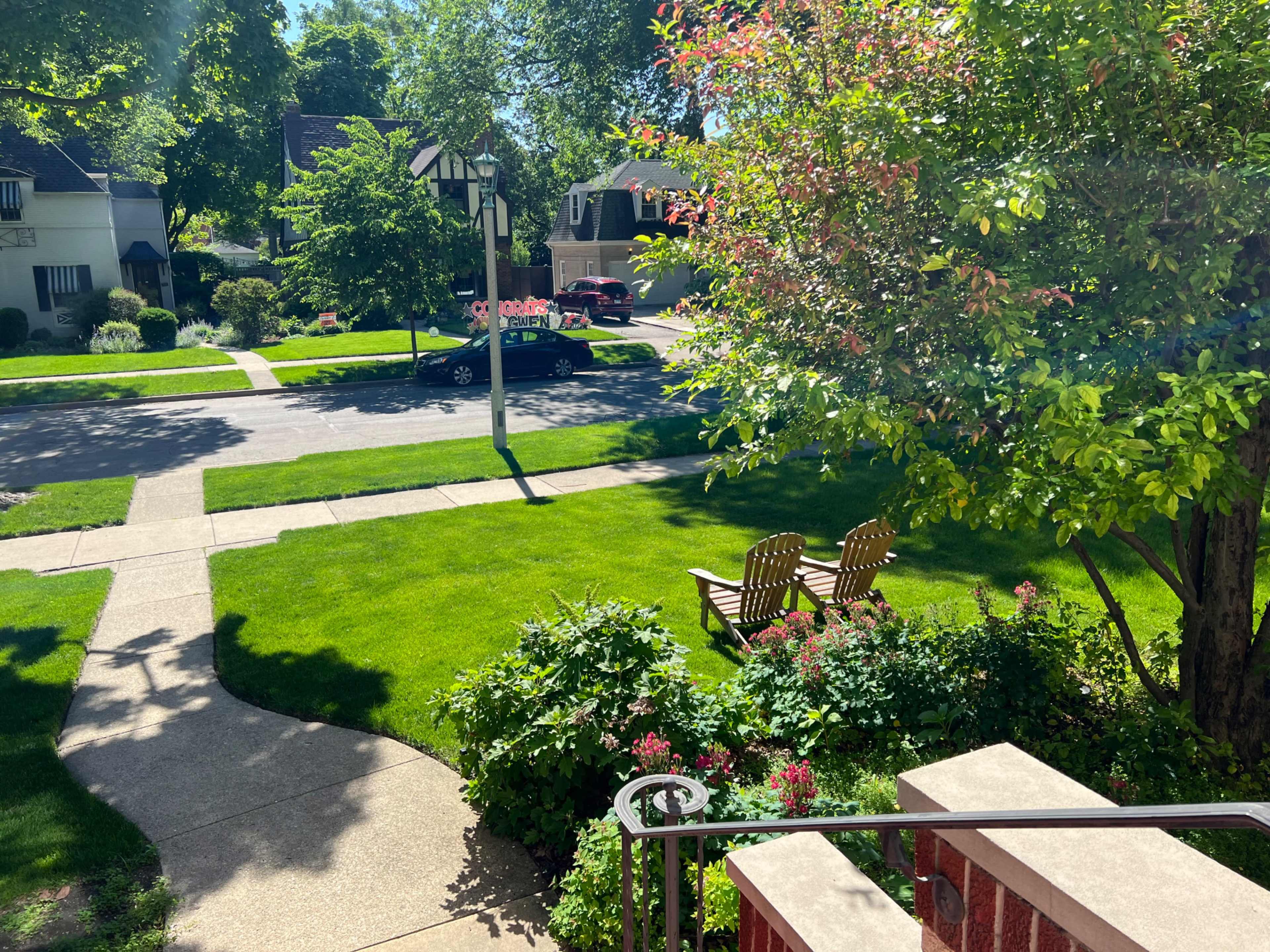 The scene shows a suburban front yard with a pathway leading to a street lined with trees and parked cars, featuring a pair of wooden chairs on the grass.