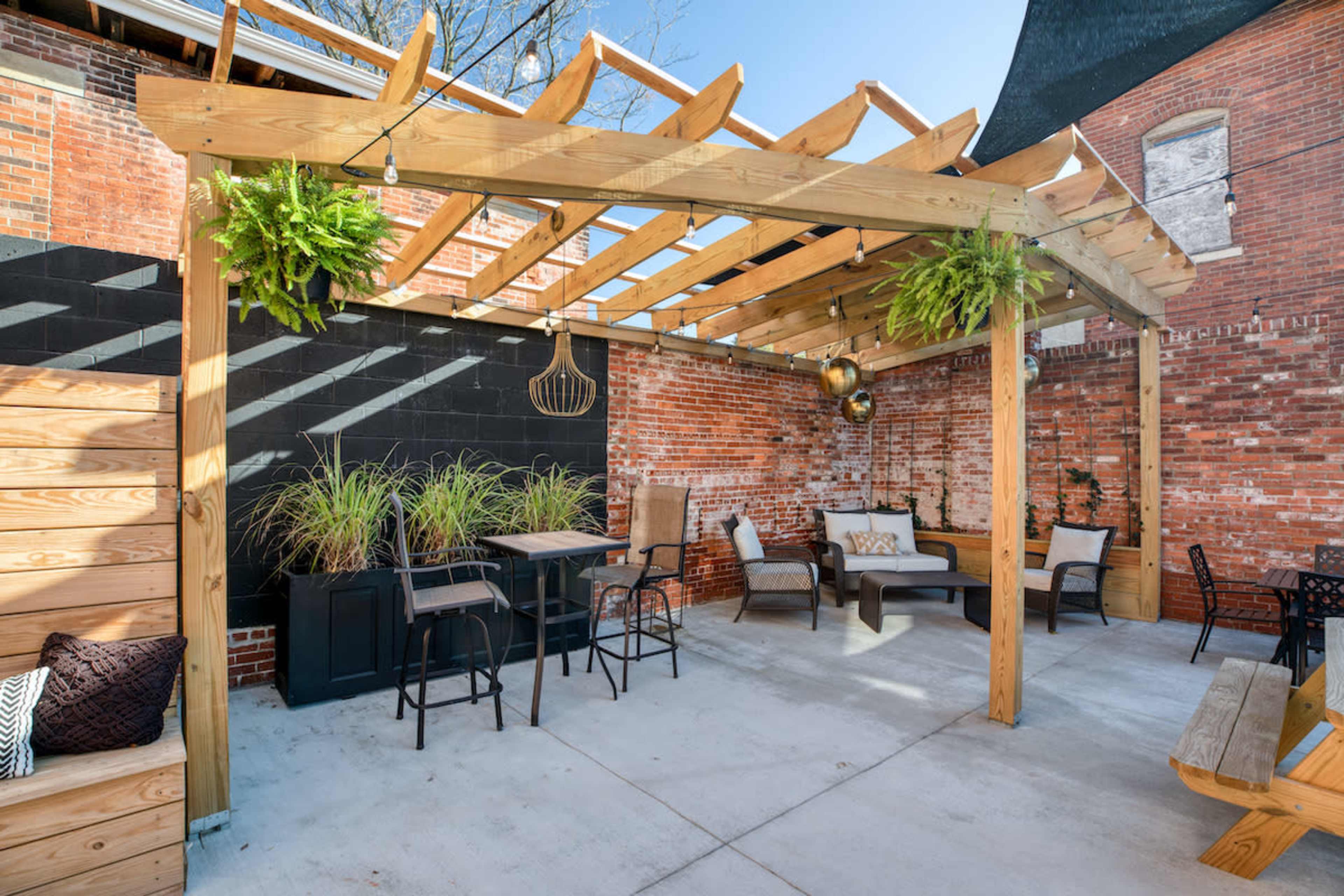 A patio area with wooden pergola, seating arrangements, and planters against a brick wall.