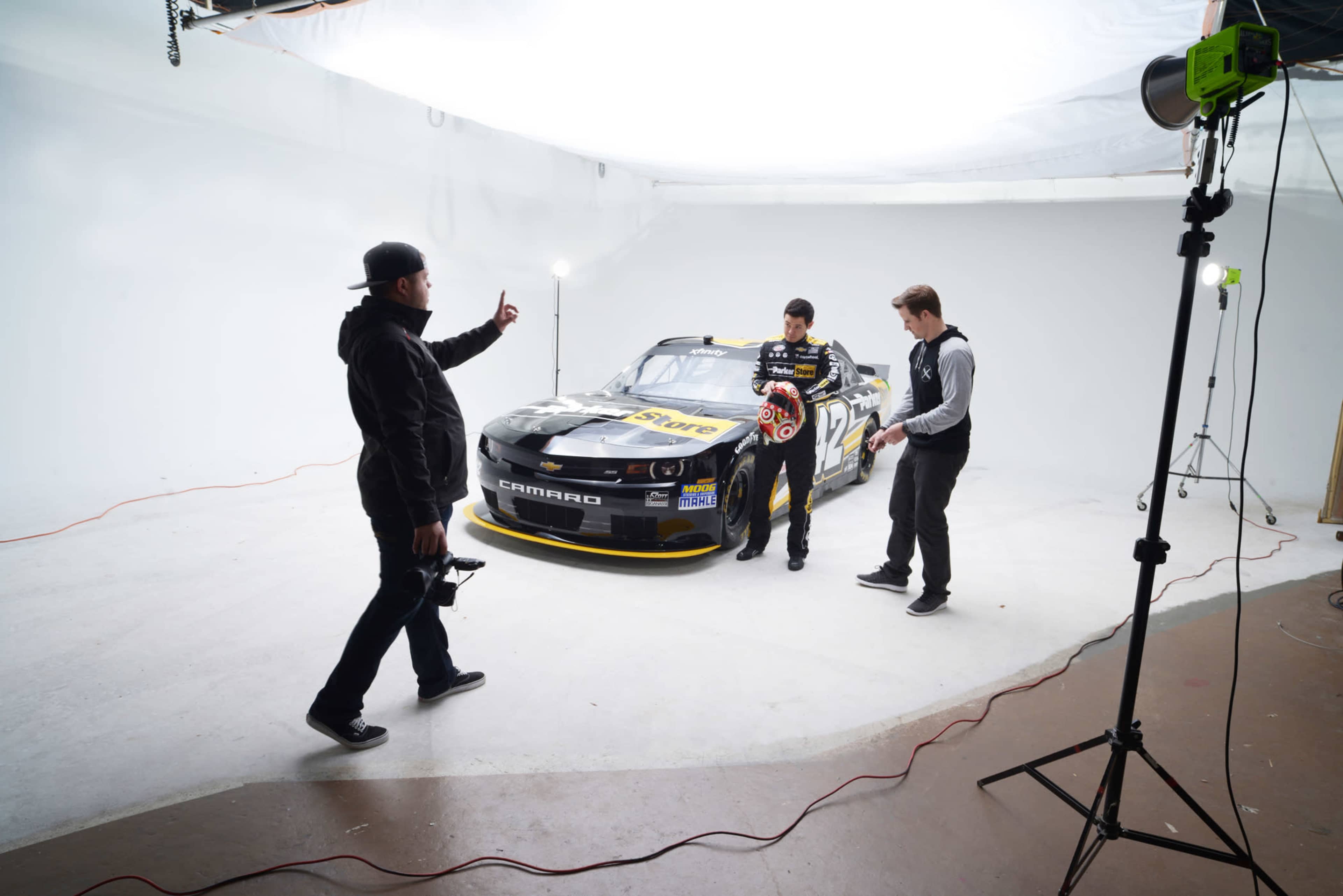 A car model is positioned in a studio while two individuals, one holding a helmet, interact with a photographer.