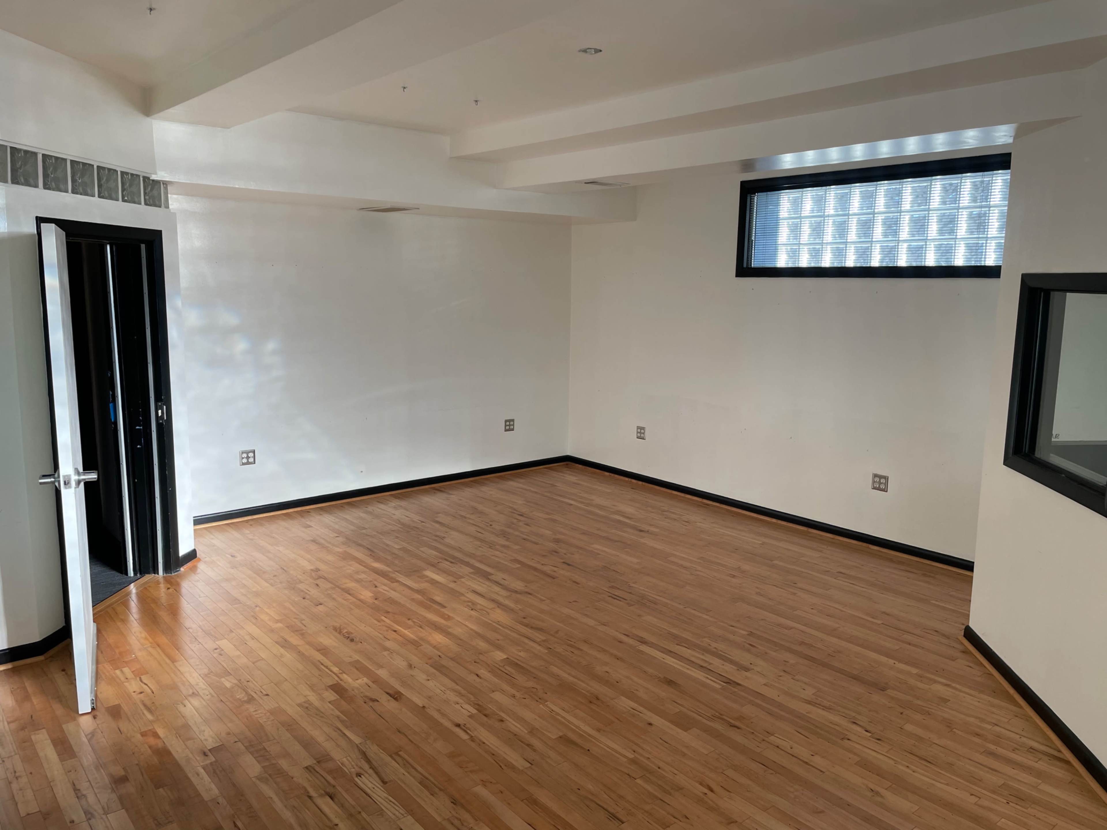 The image shows a vacant room with hardwood flooring, white walls, and large windows, featuring an open door on one side.