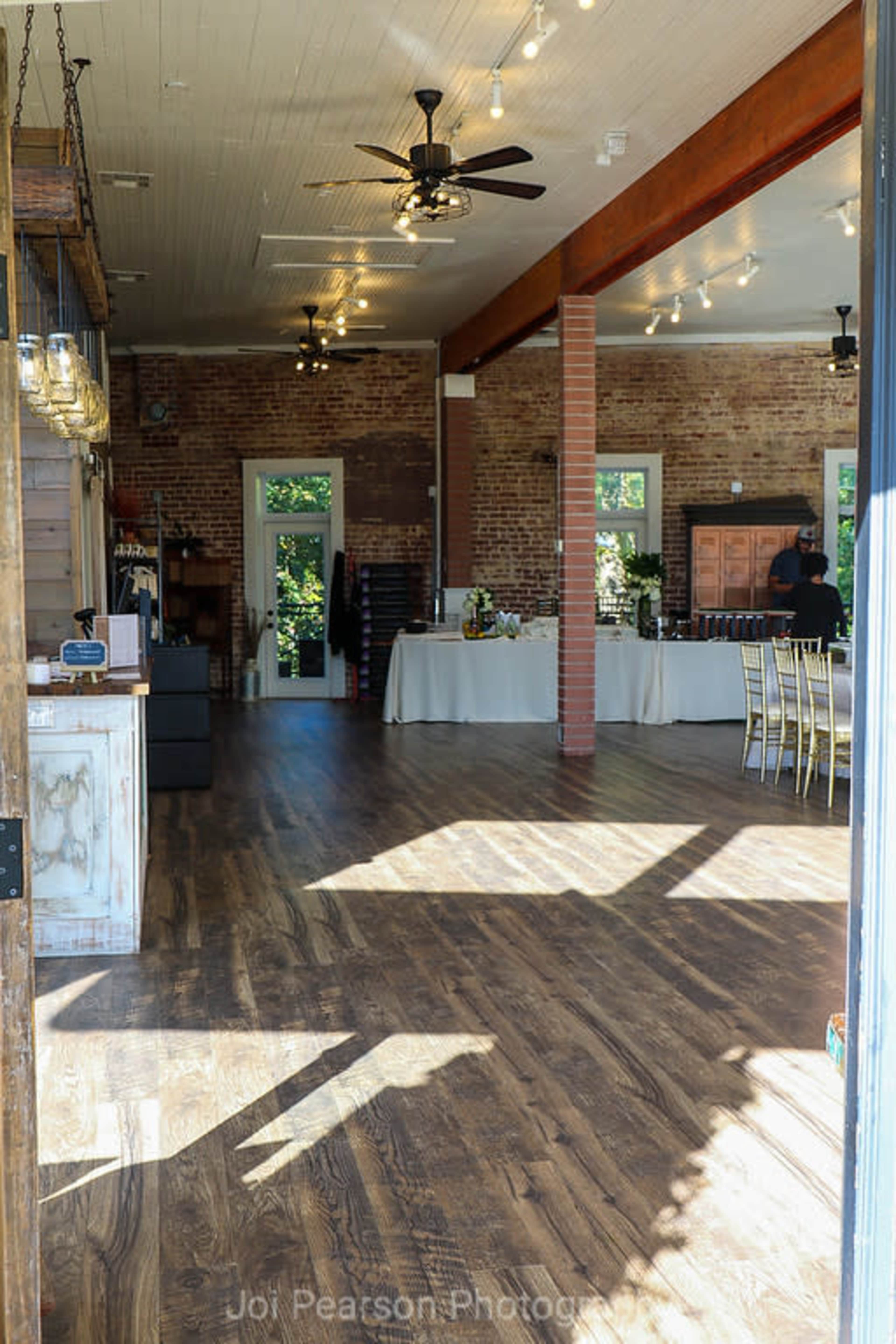 A spacious, well-lit interior of an event venue with wooden flooring, exposed brick walls, and various tables set up for an occasion.