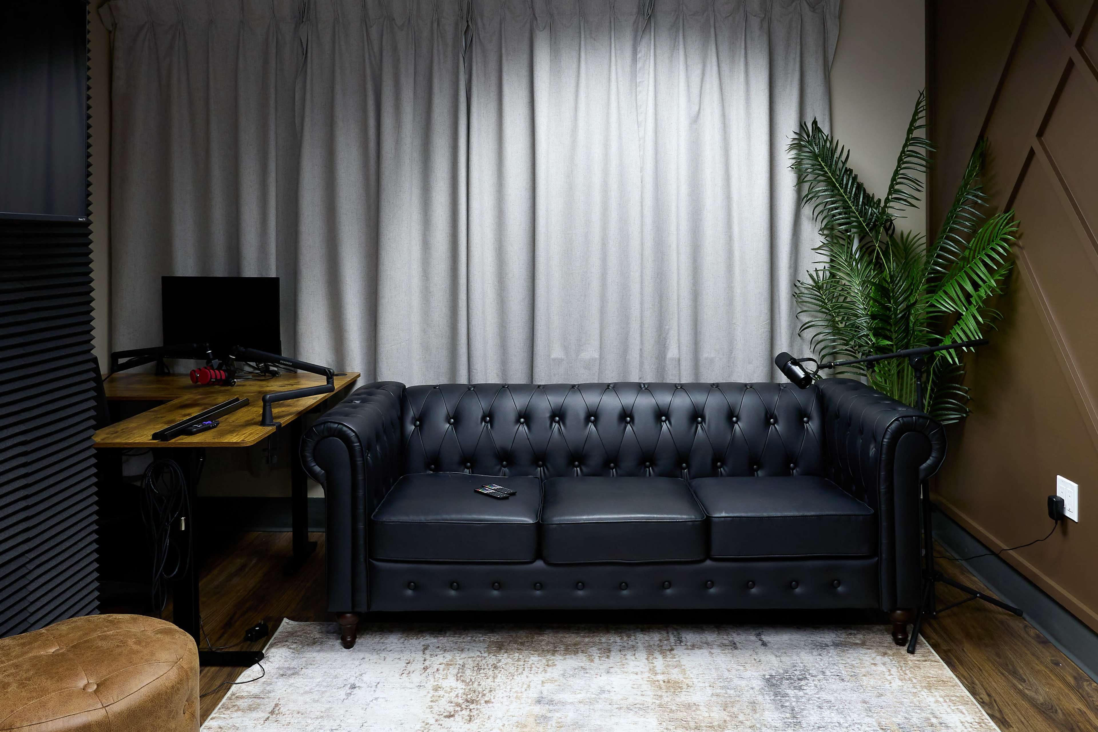 The image shows a black leather tufted sofa in front of a window with light gray curtains, accompanied by a small wooden desk with a computer monitor and a decorative plant nearby.