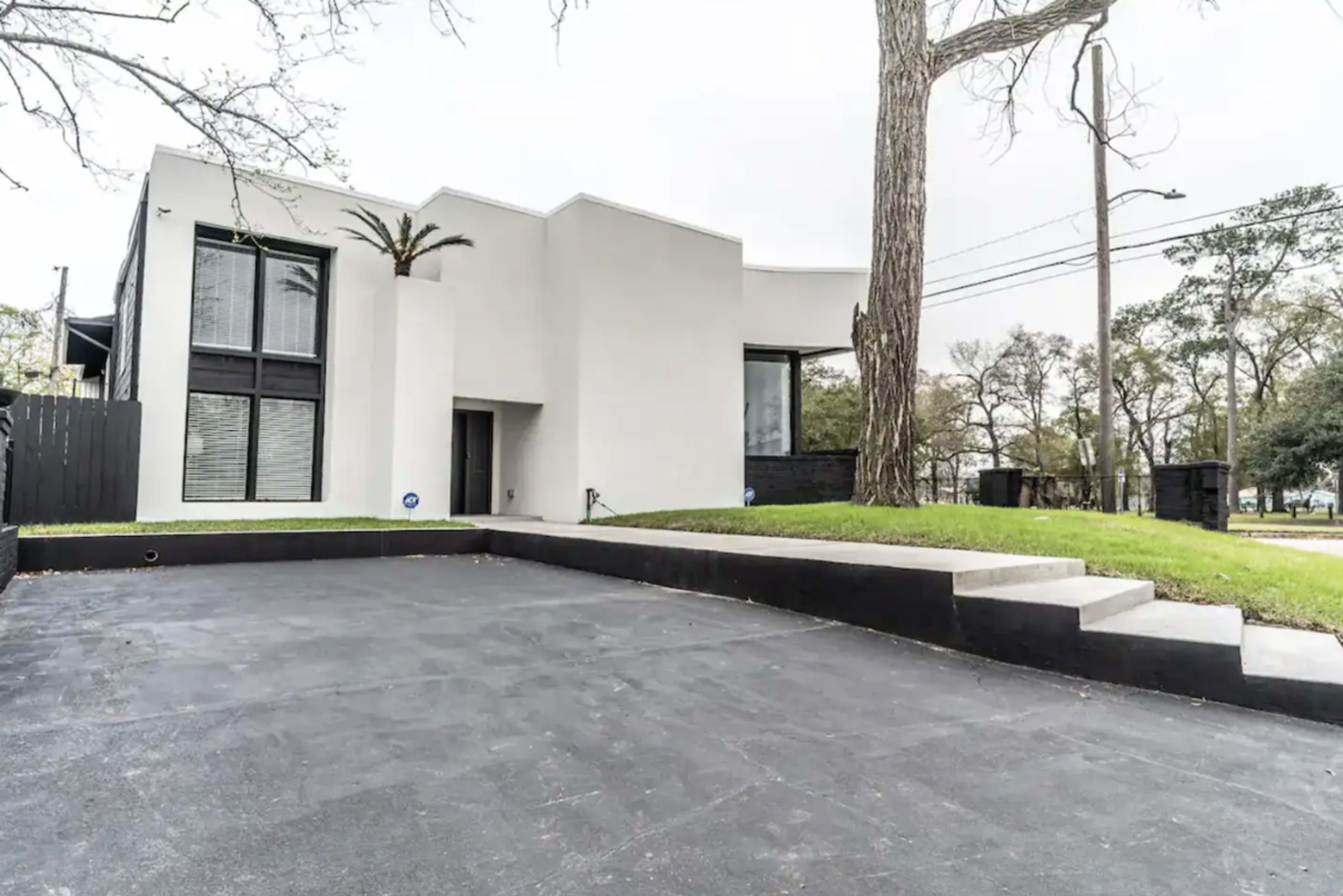 A modern, minimalist white house with large windows and a flat roof, surrounded by green grass and trees, with a concrete driveway leading to the entrance.
