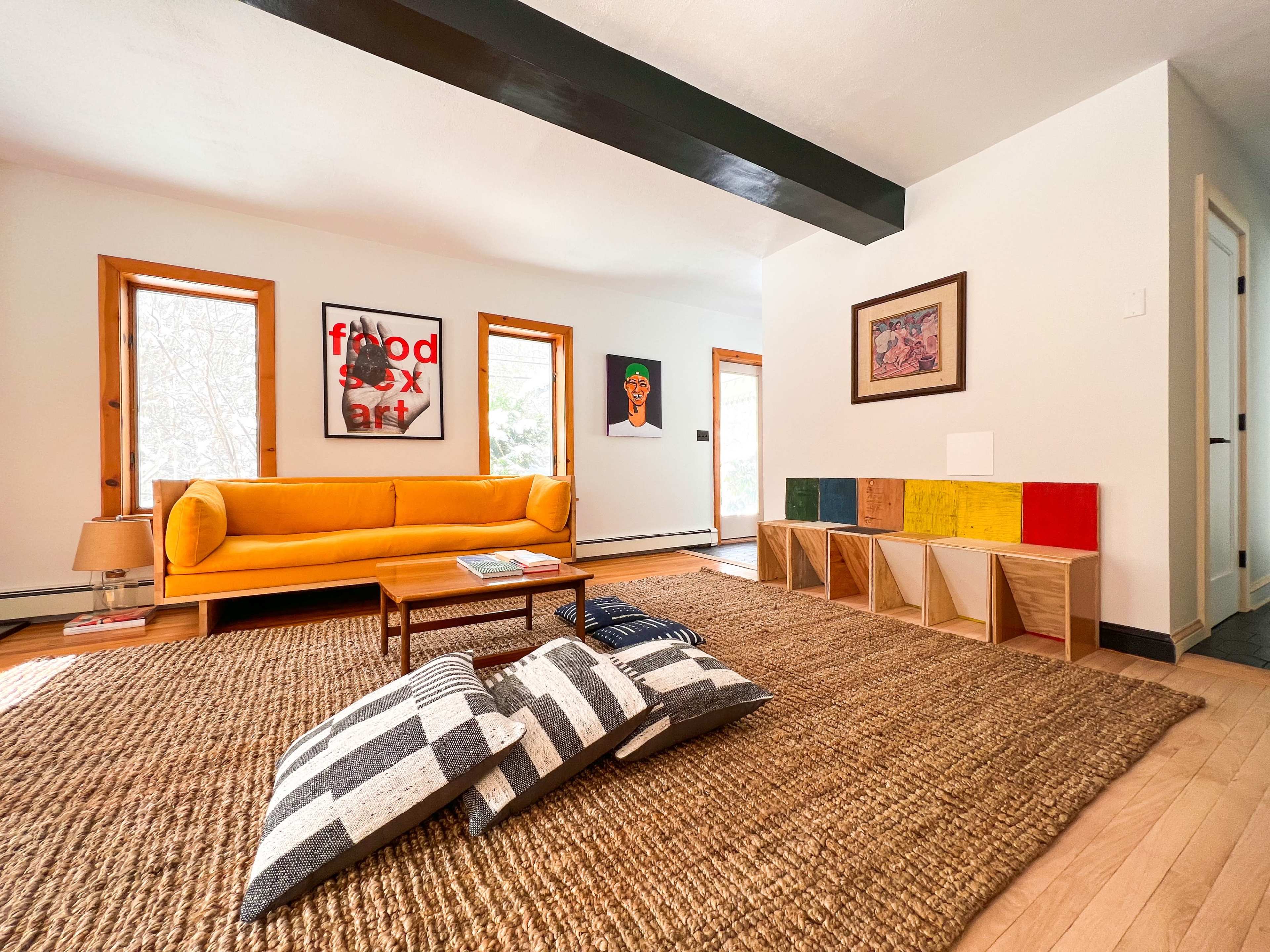 The living room features a bright orange sofa, a wooden coffee table, and a large woven rug, accompanied by colorful wall art and a series of storage cubes.
