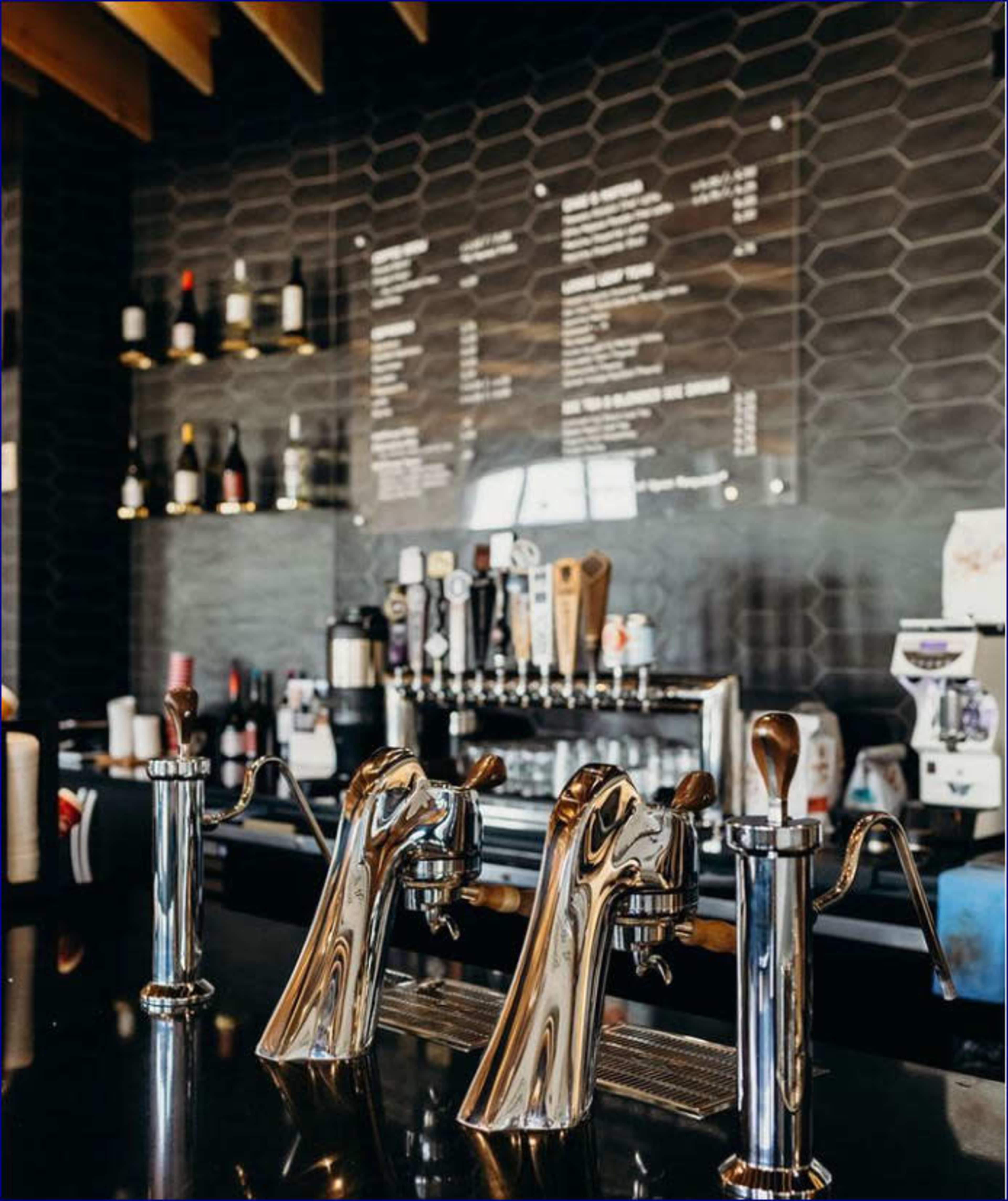 The image shows a bar area with three shiny beer taps in the foreground, a menu displayed on the wall behind them, and various bottles and equipment on shelves.