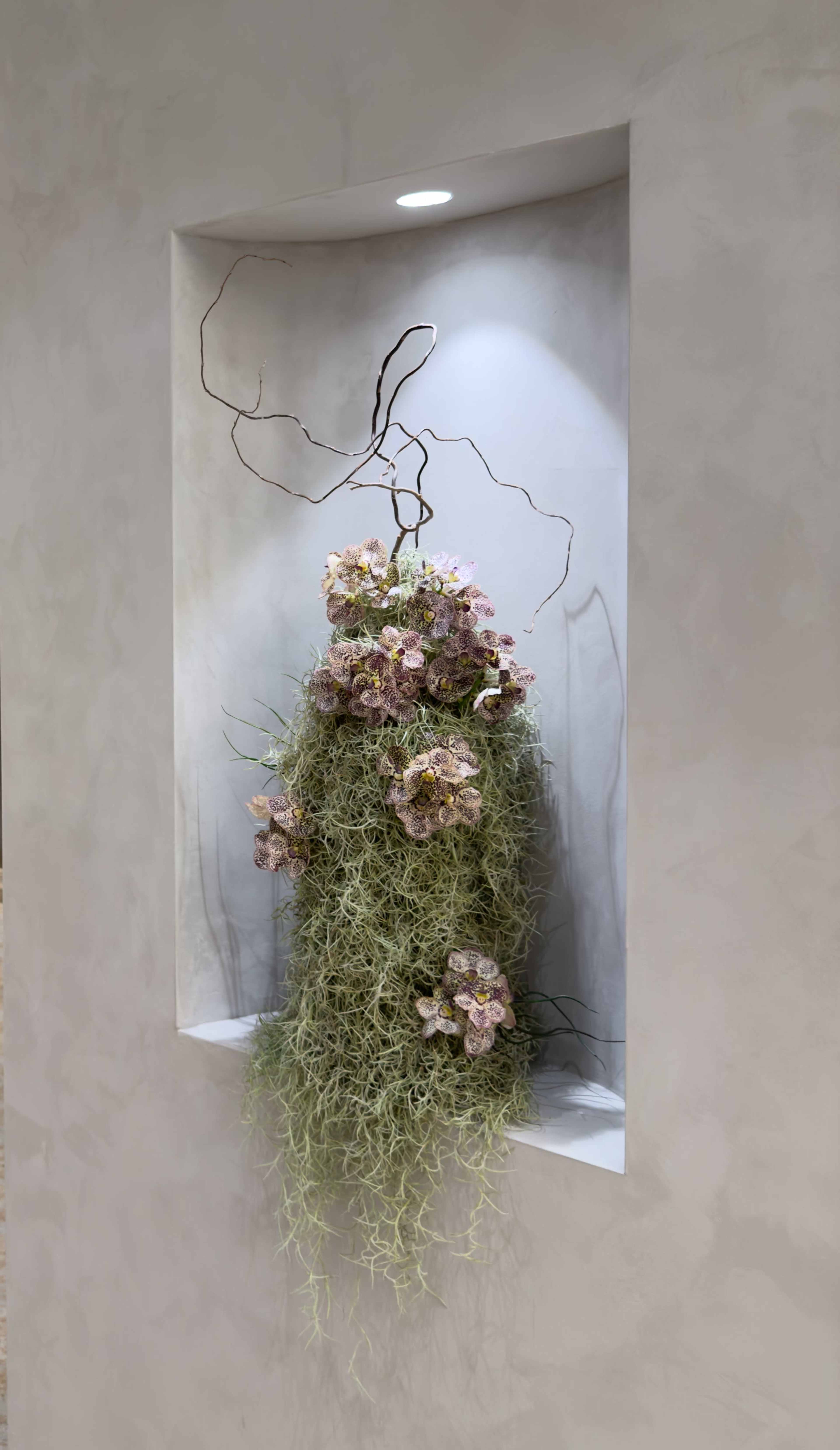 A wall-mounted floral arrangement features a cluster of dried flowers and Spanish moss set against a textured background.