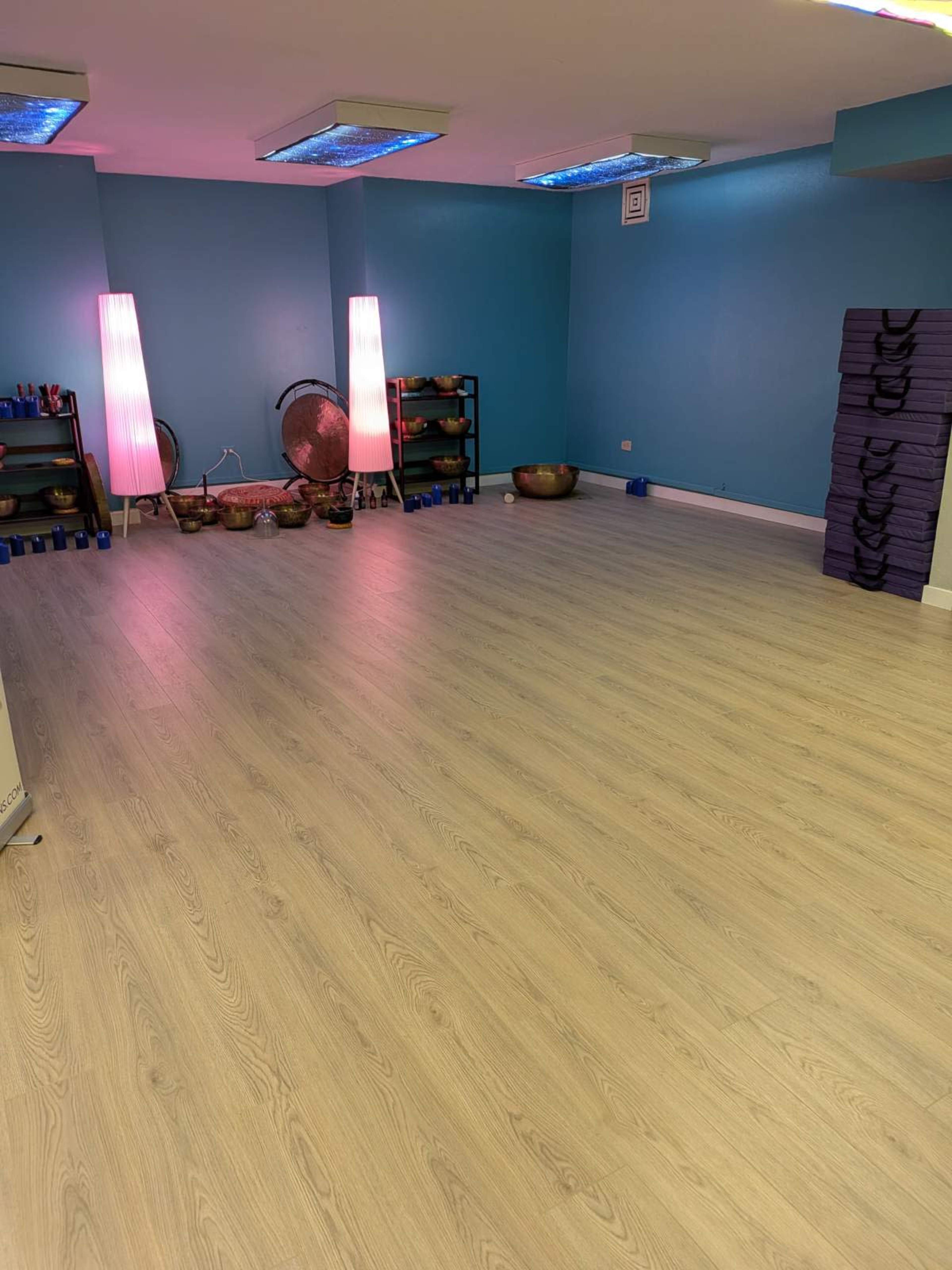 The image shows a spacious room with light-colored wooden flooring, blue walls, dim lighting, and various items for relaxation including storage shelves and yoga mats.