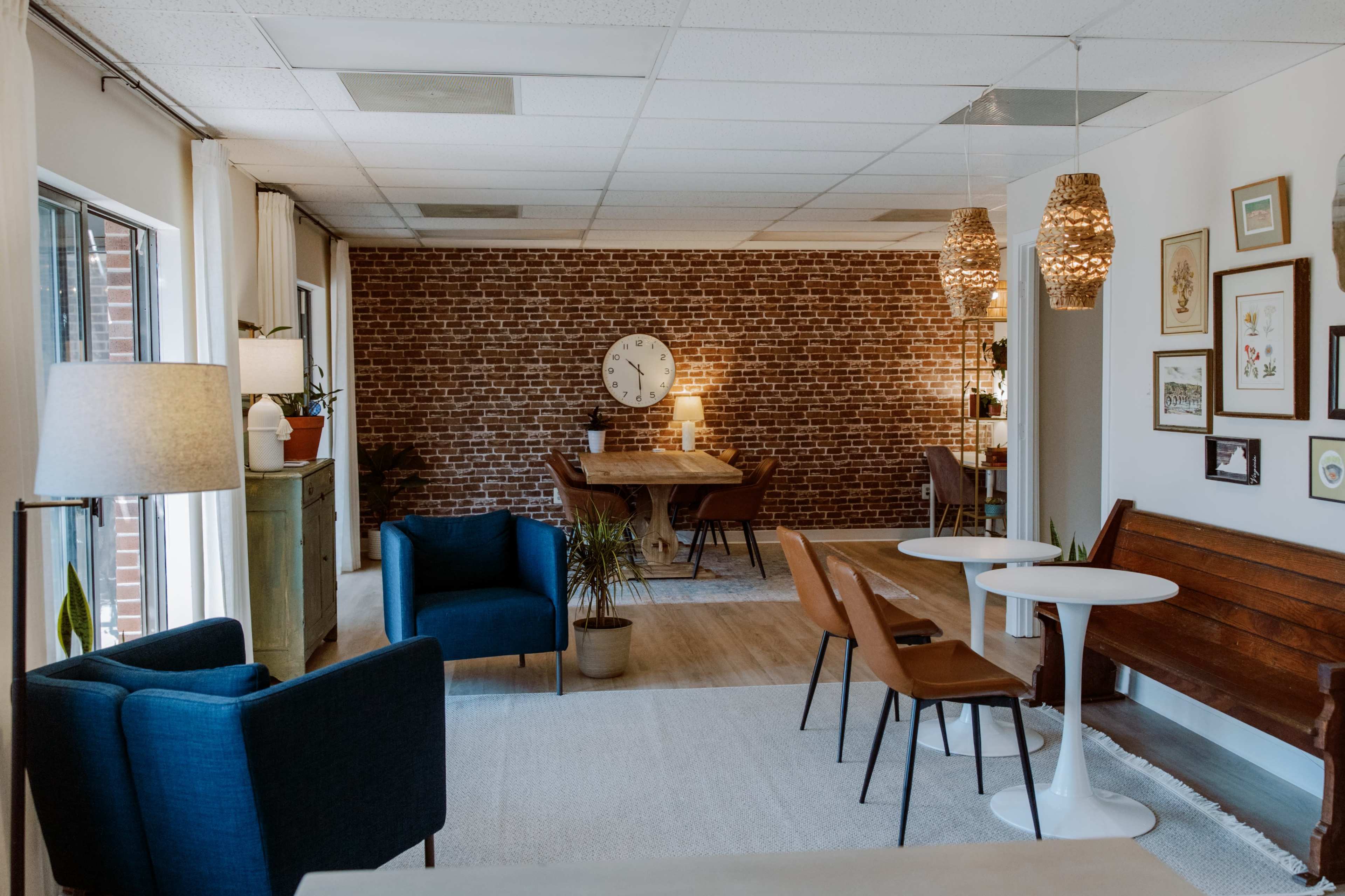 The image shows a modern interior space featuring a seating area with two blue chairs and a wooden table surrounded by brick walls, with a dining area in the background that includes a clock and wall art.