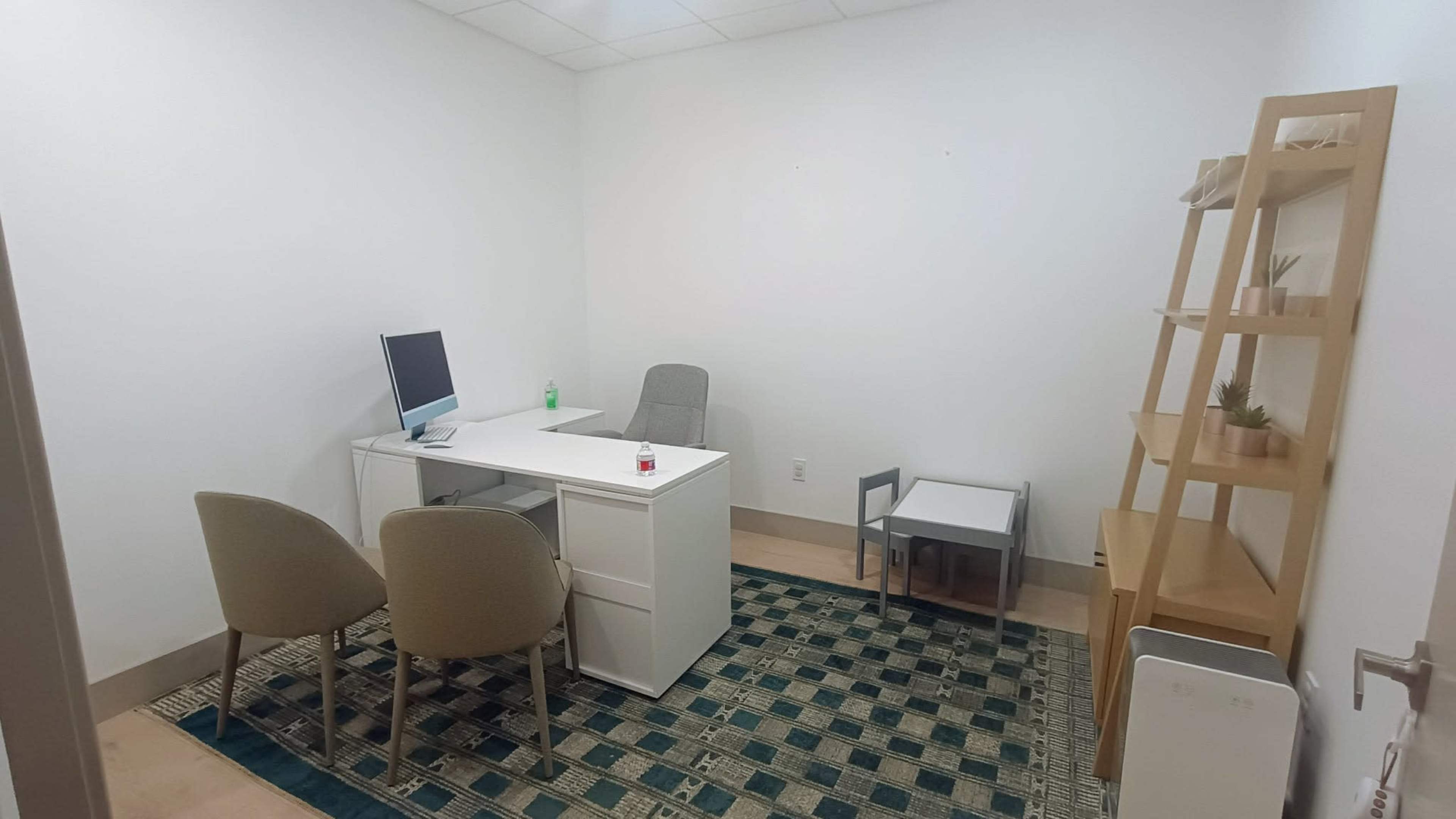 A small, minimalist office features a desk with a computer, two chairs, a small side table, and a bookshelf against one wall.