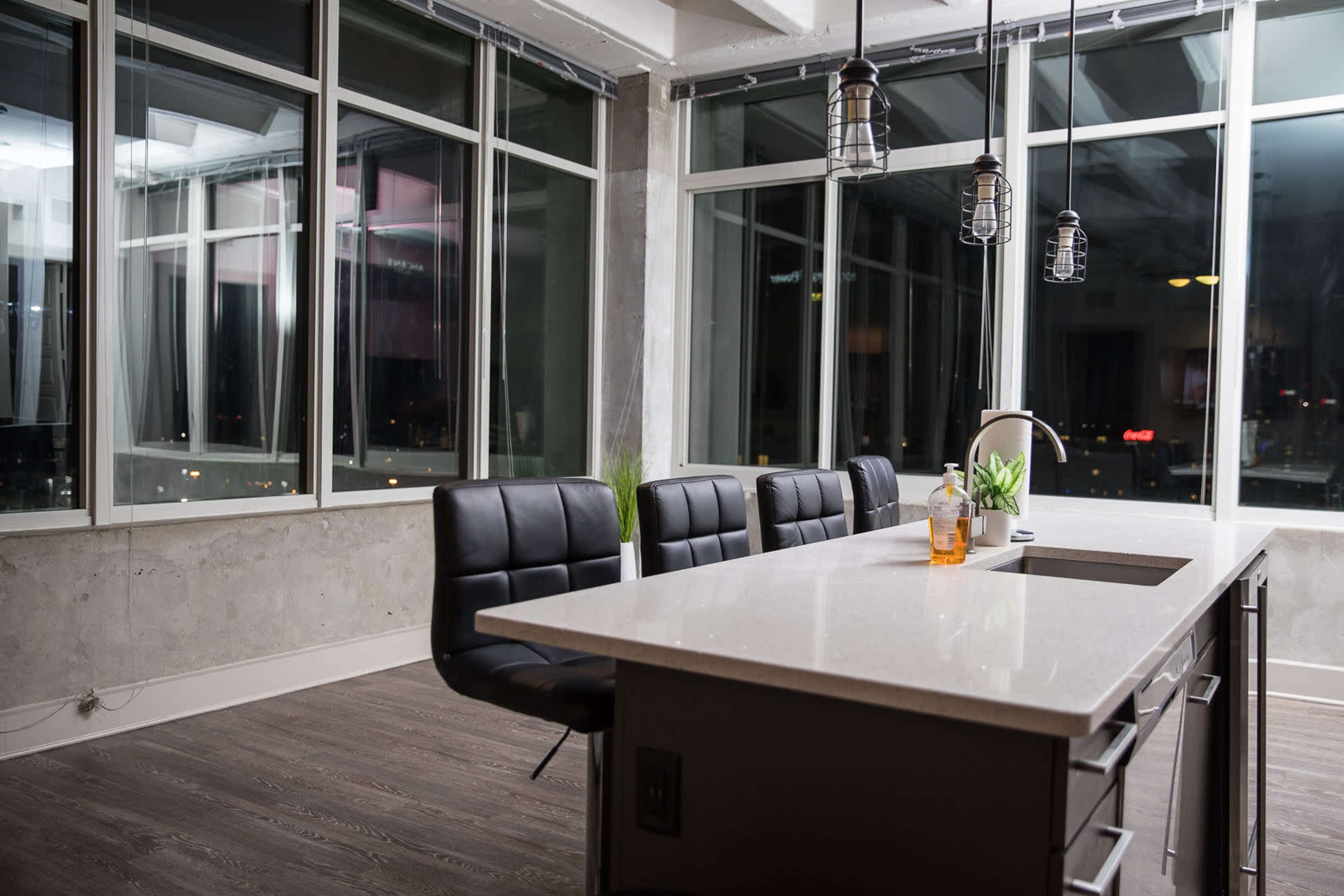 A modern kitchen features a central island with a white countertop, surrounded by black leather chairs and large windows offering a view of the city.