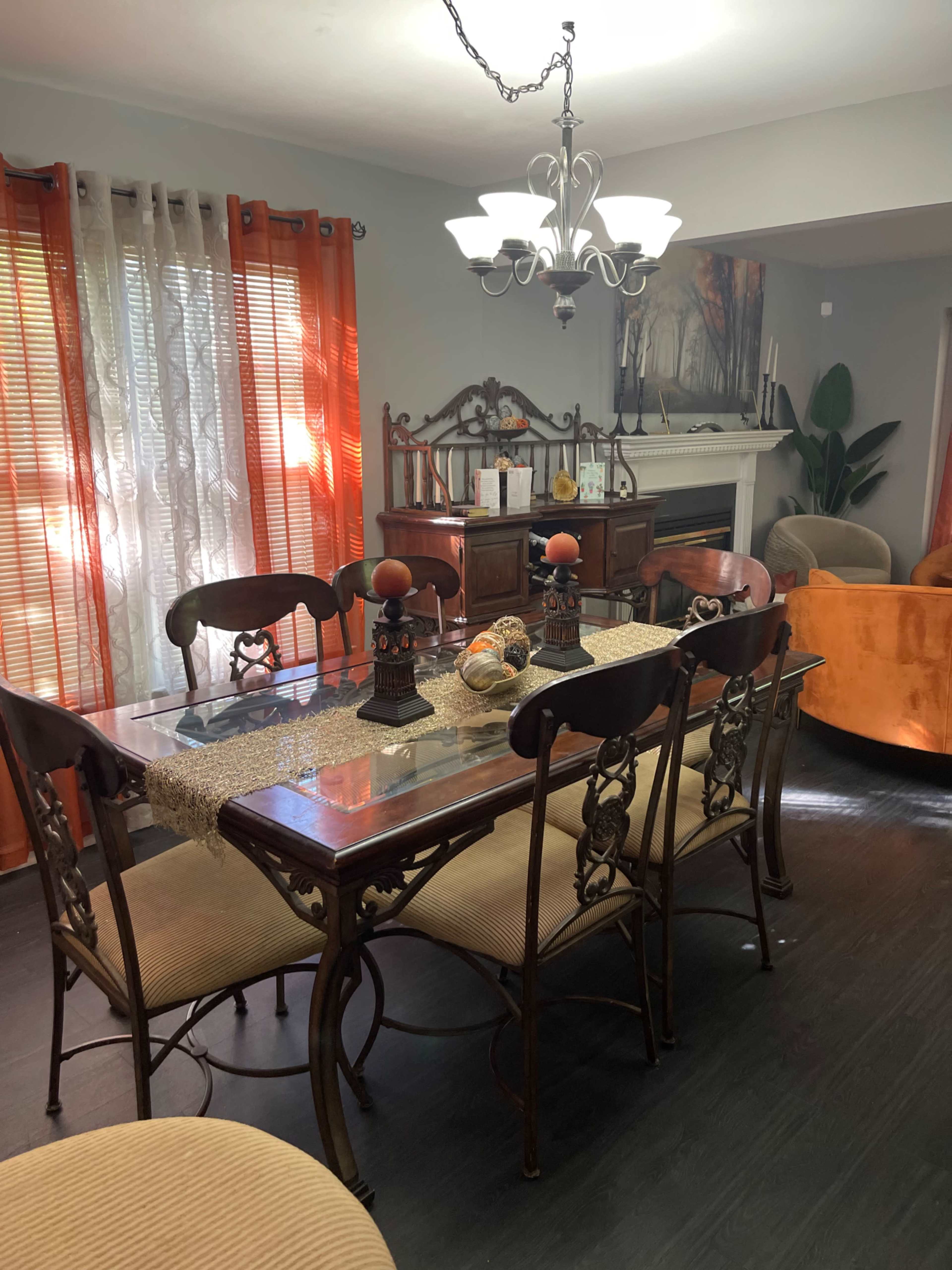 The image shows a dining area with a wooden table surrounded by chairs, a decorative centerpiece, and large windows with sheer orange curtains.