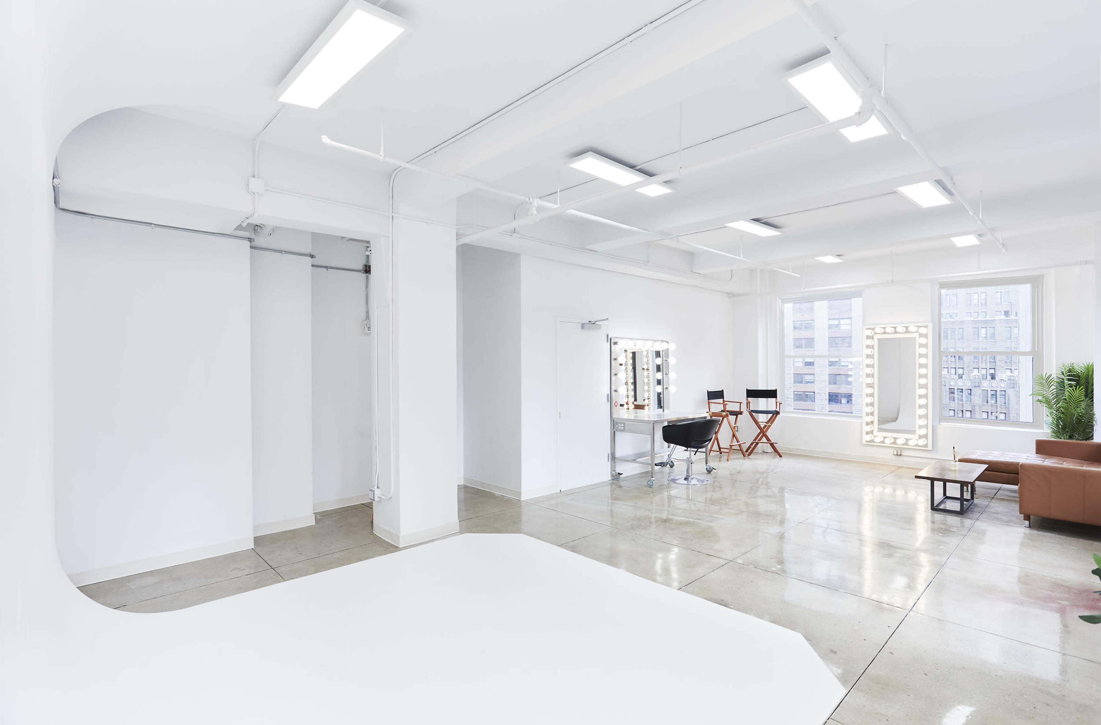 The image shows a spacious, minimalist studio with a bright white backdrop, a vanity with lights, two director's chairs, and a seating area with a brown couch.
