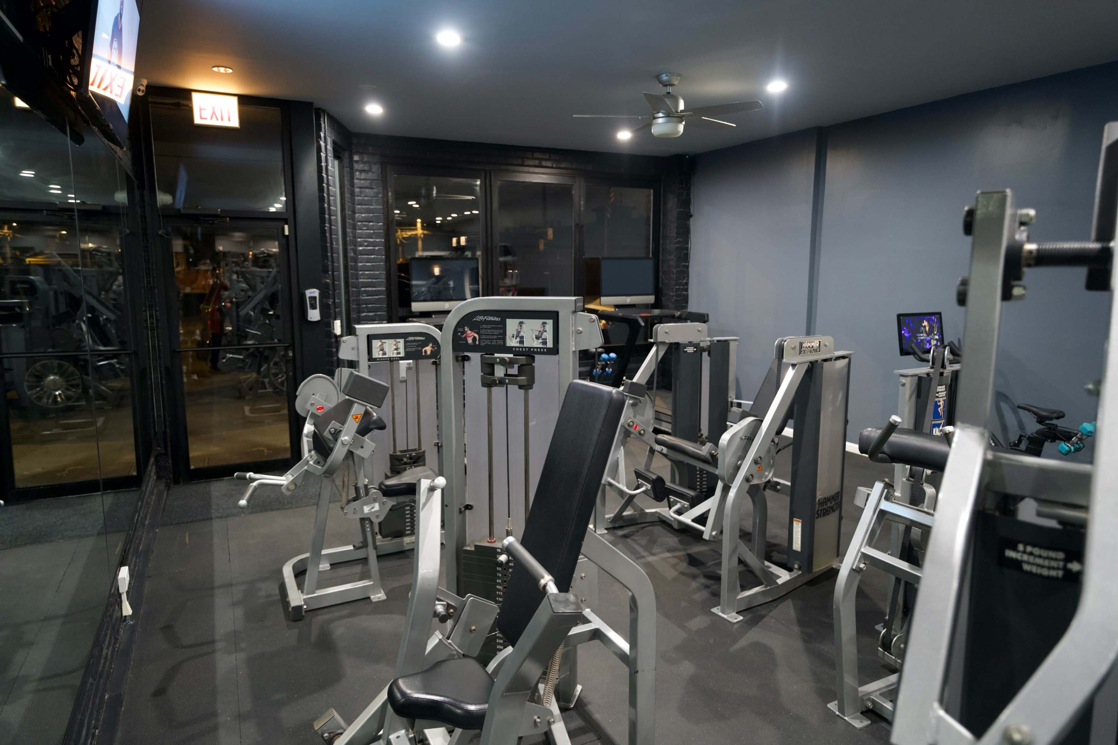 A fitness gym with various exercise machines arranged in an indoor space featuring dark walls and ceiling lights.