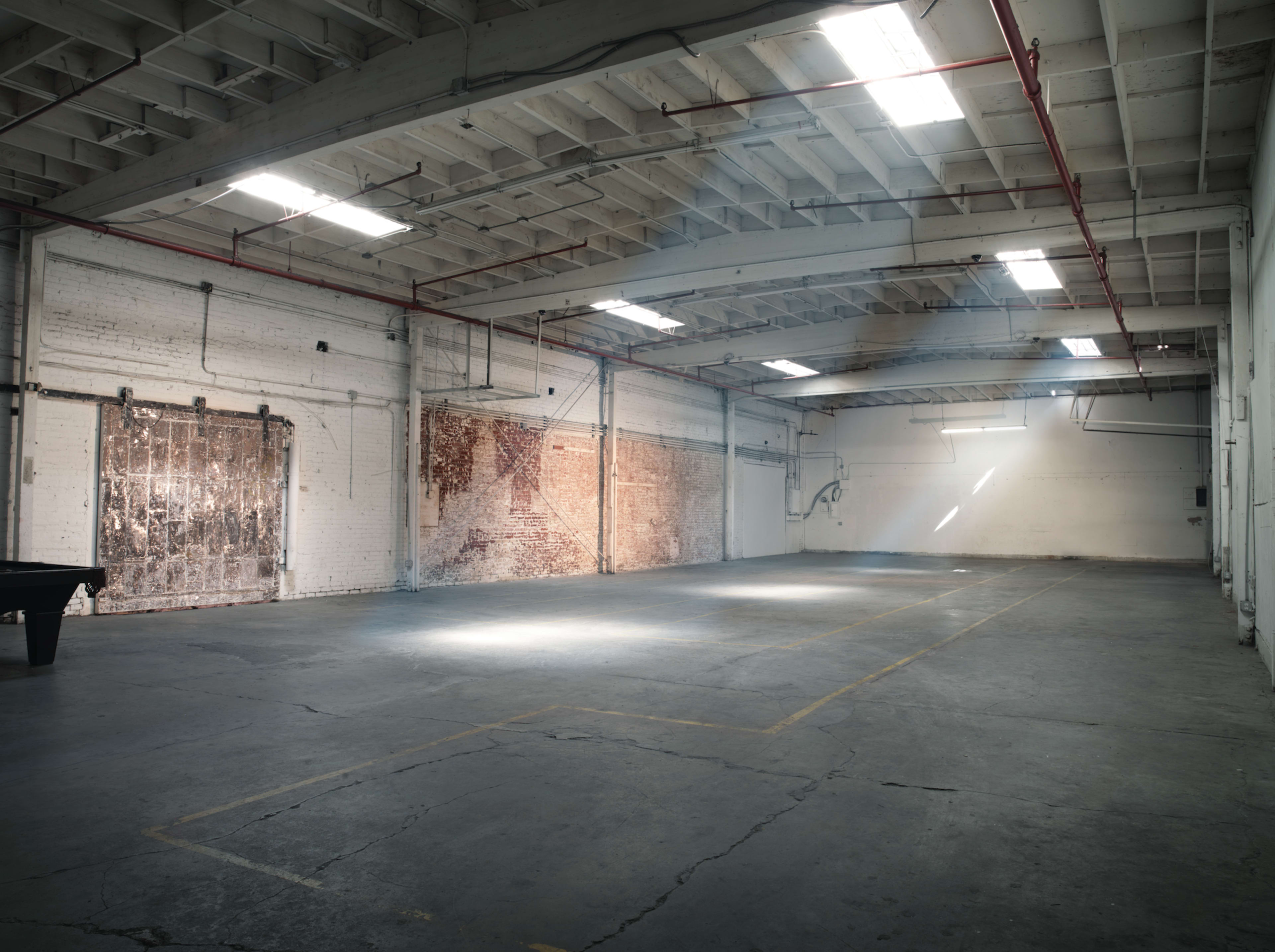 An empty industrial space features large windows with light beams illuminating the concrete floor and brick walls.