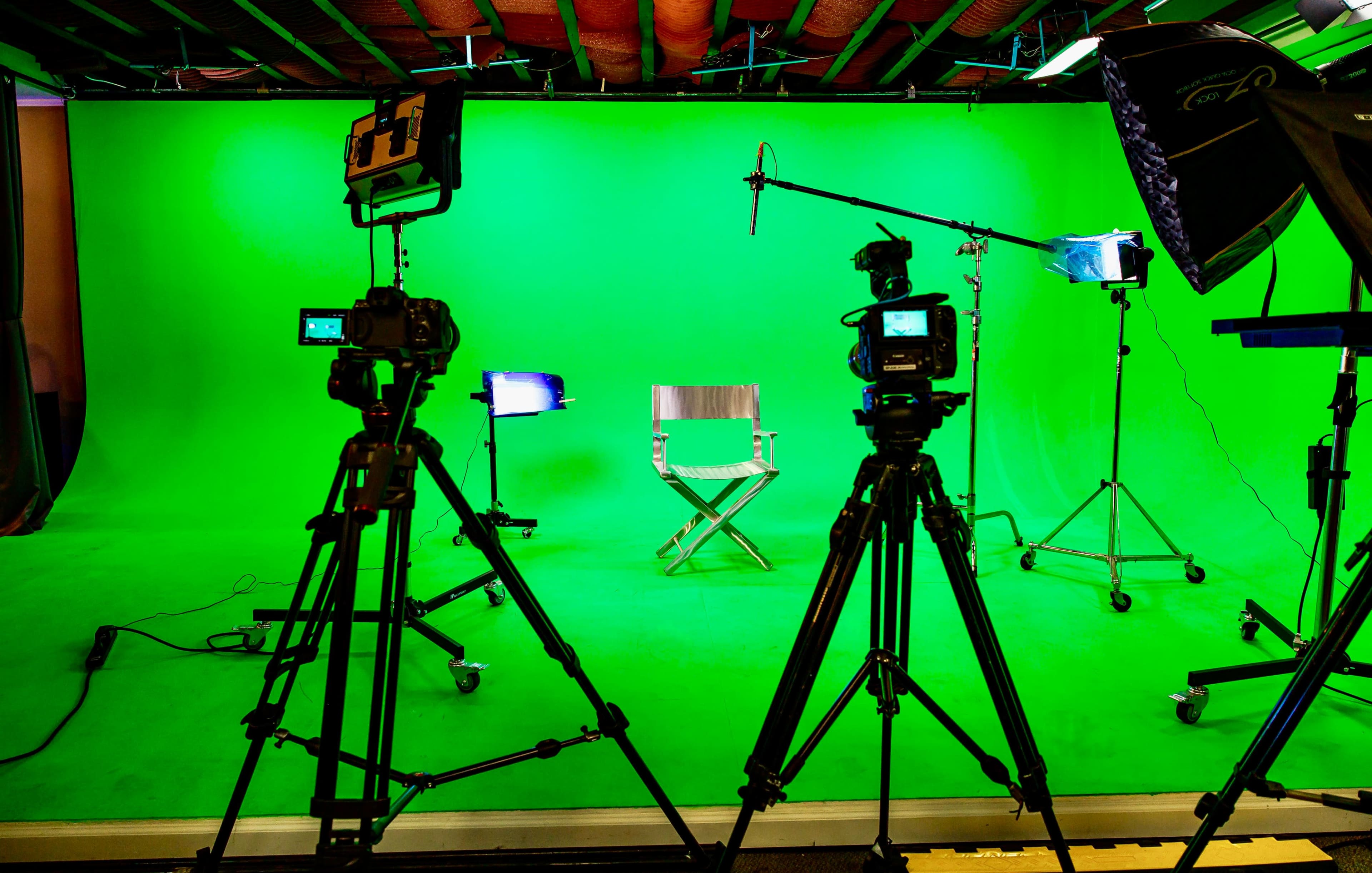 A television production set features a green backdrop, a director's chair in the center, and multiple cameras and lights positioned around it.