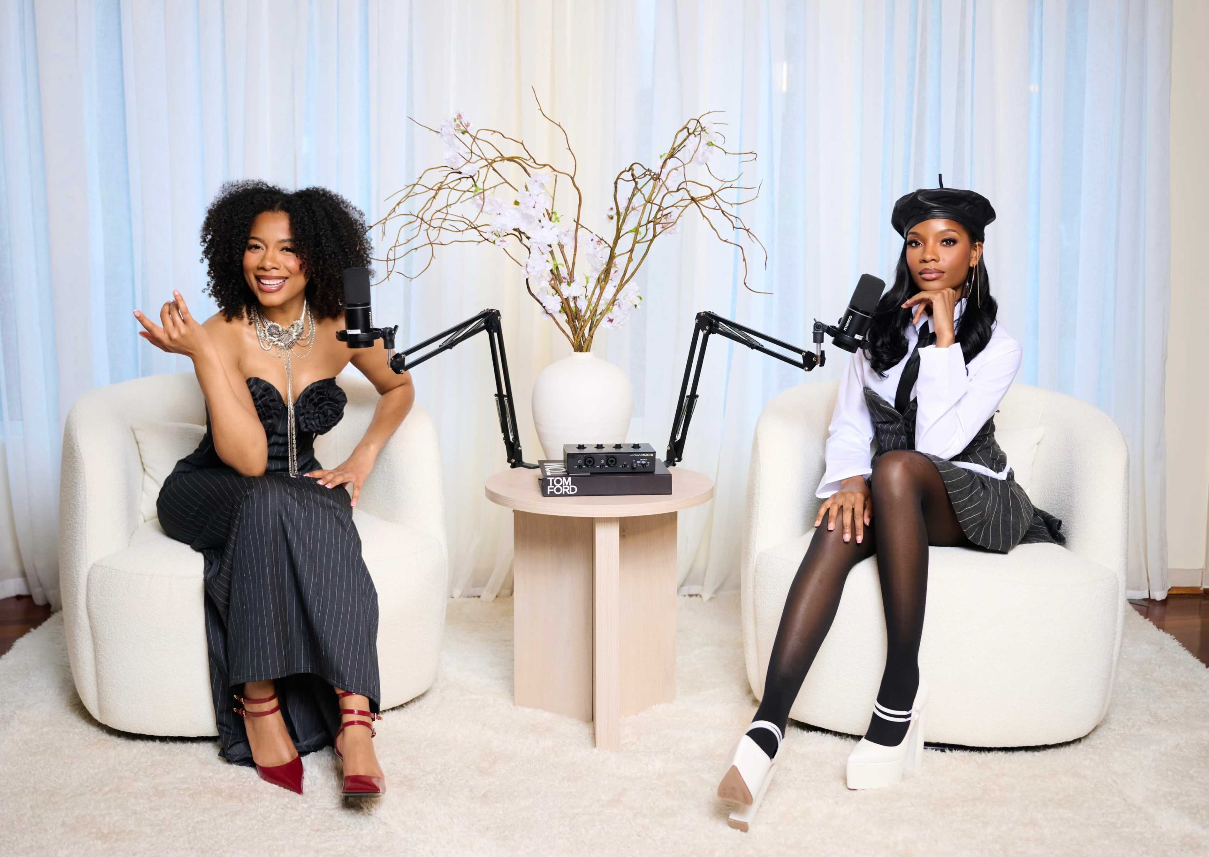 Two women are seated on plush chairs in a light-filled room, each positioned in front of a microphone and a small table adorned with a floral arrangement.