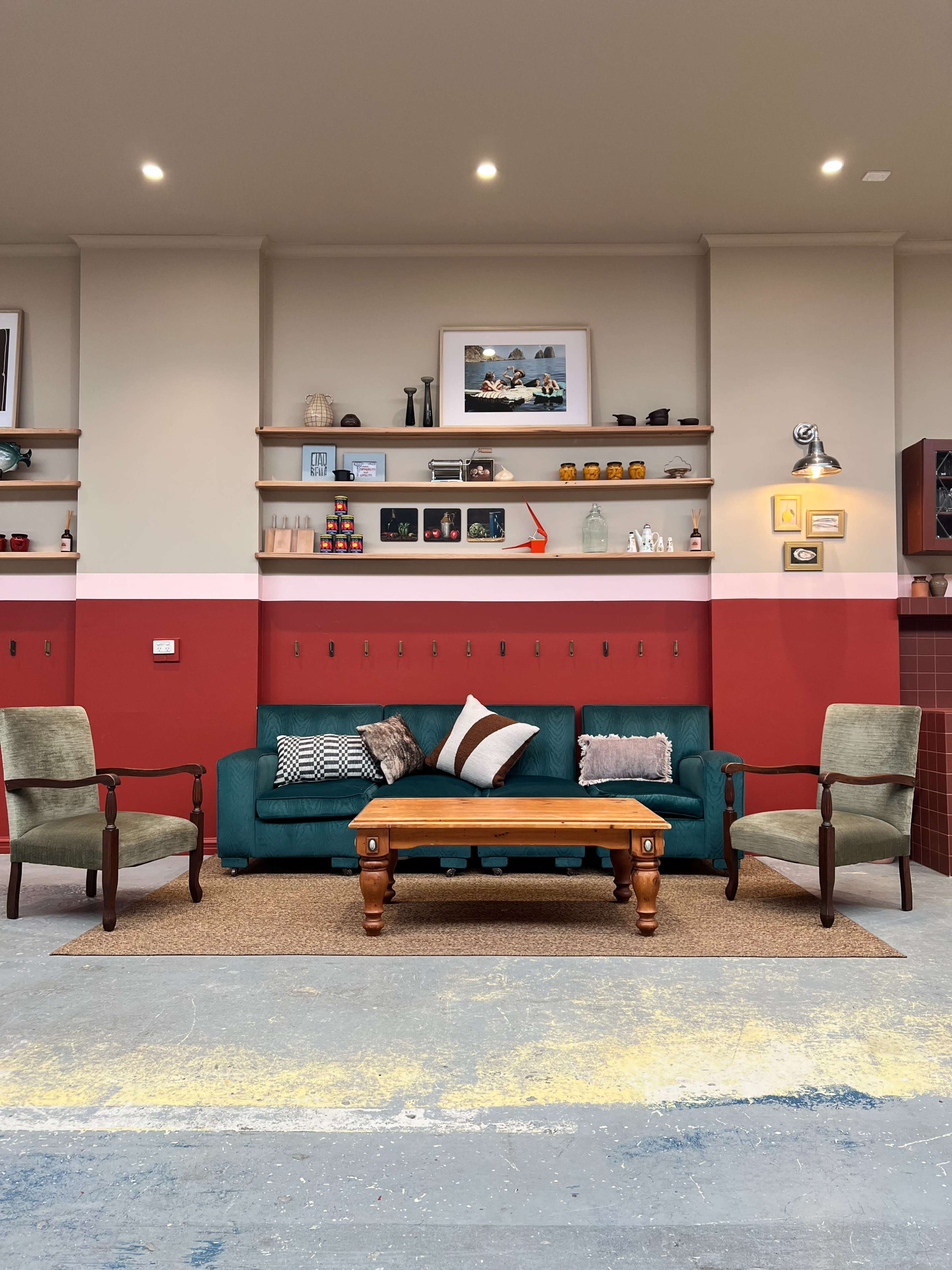 The image shows a cozy living room setup with a green couch, two armchairs, and a wooden coffee table against a backdrop of red and white walls adorned with shelves displaying various items.