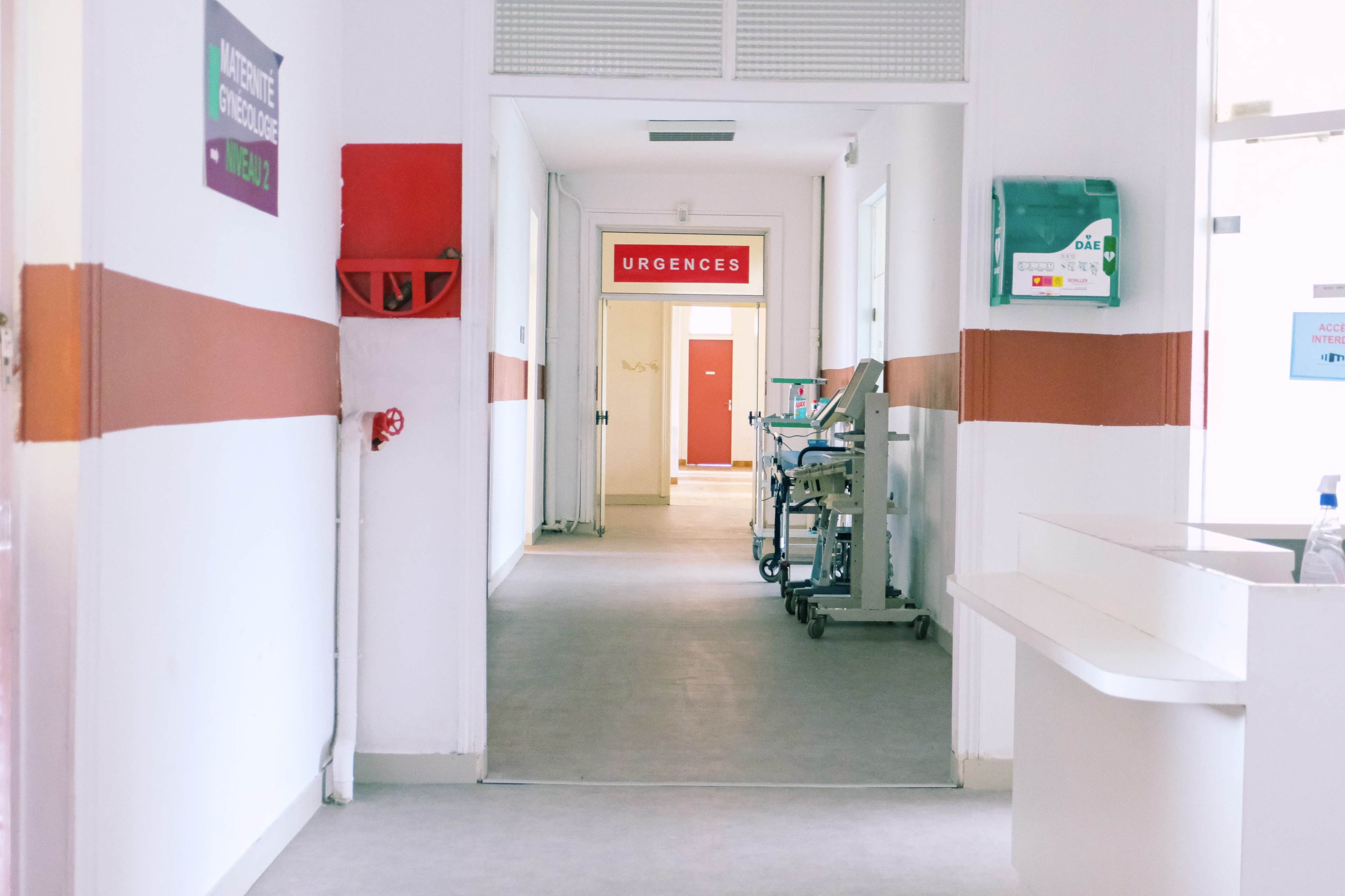 A hospital corridor with a sign indicating "Urgences" at the far end and medical equipment positioned along the wall.
