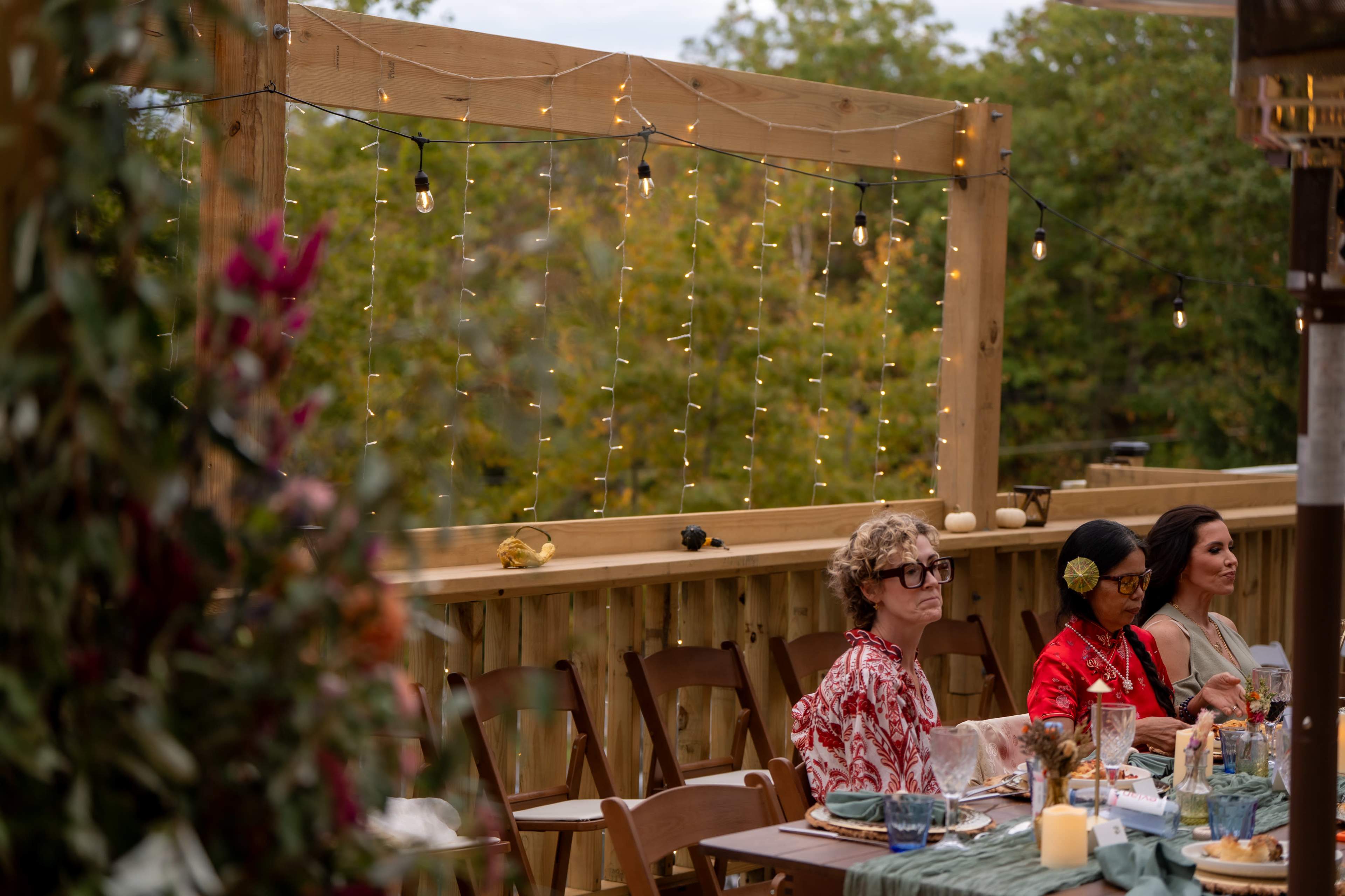 A group of three women sits at a decorated table outdoors, surrounded by string lights and greenery.