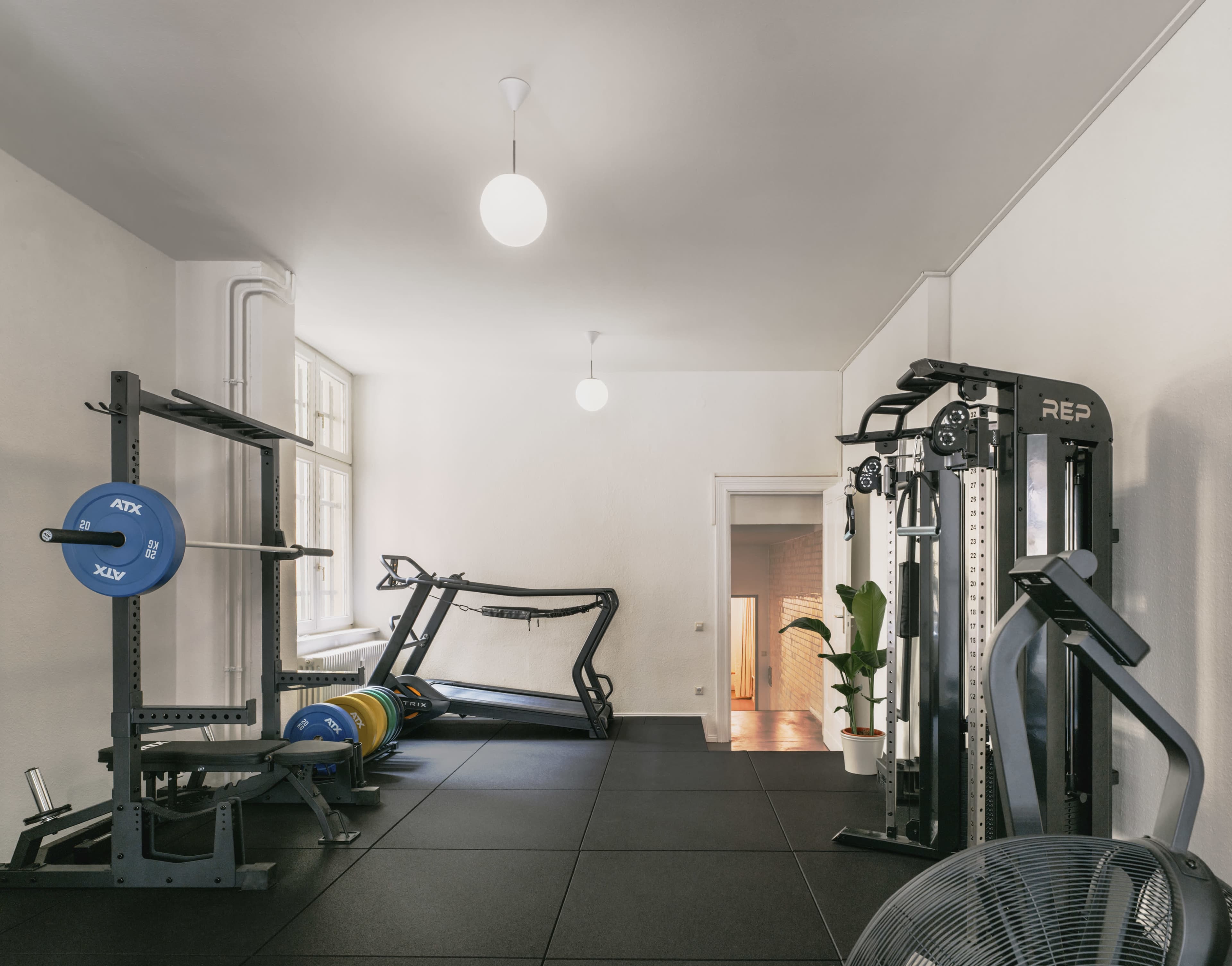The image shows a modern gym area with various exercise equipment, including weights, a treadmill, and a cable machine, situated on black rubber flooring.