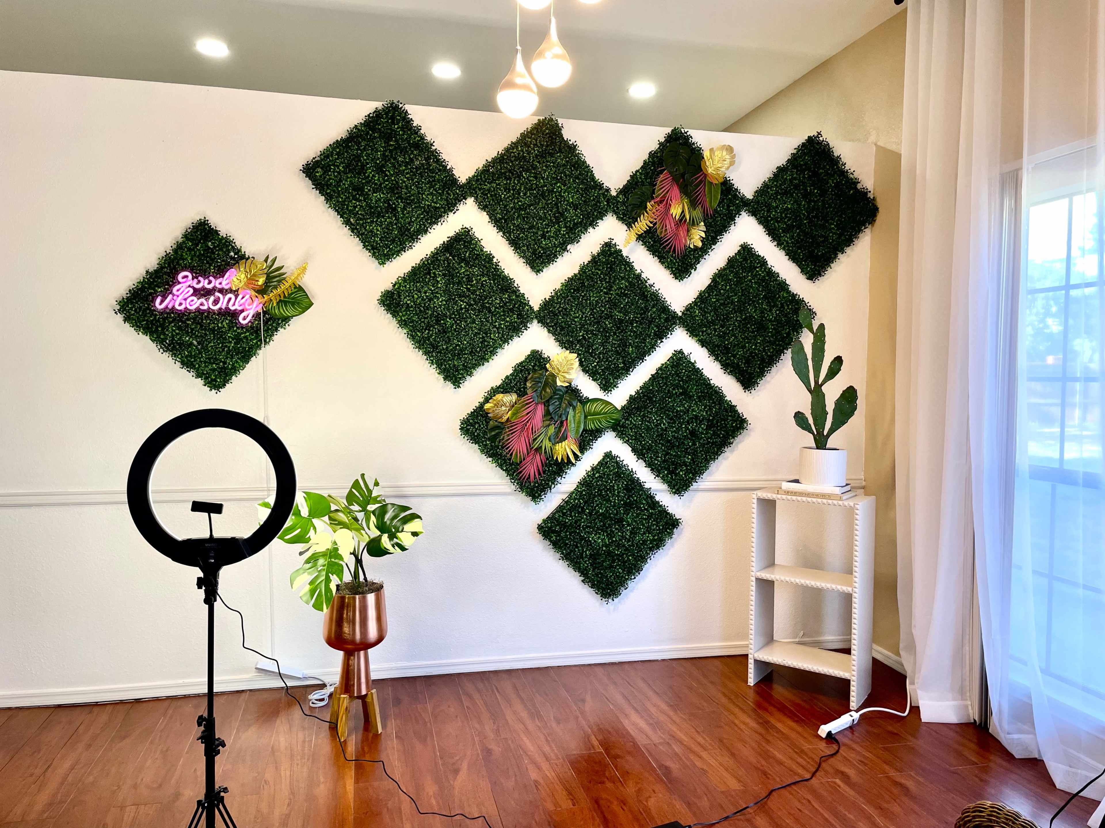The image shows a room with a decorative wall of artificial greenery arranged in diamond shapes, complemented by a neon sign and potted plants.