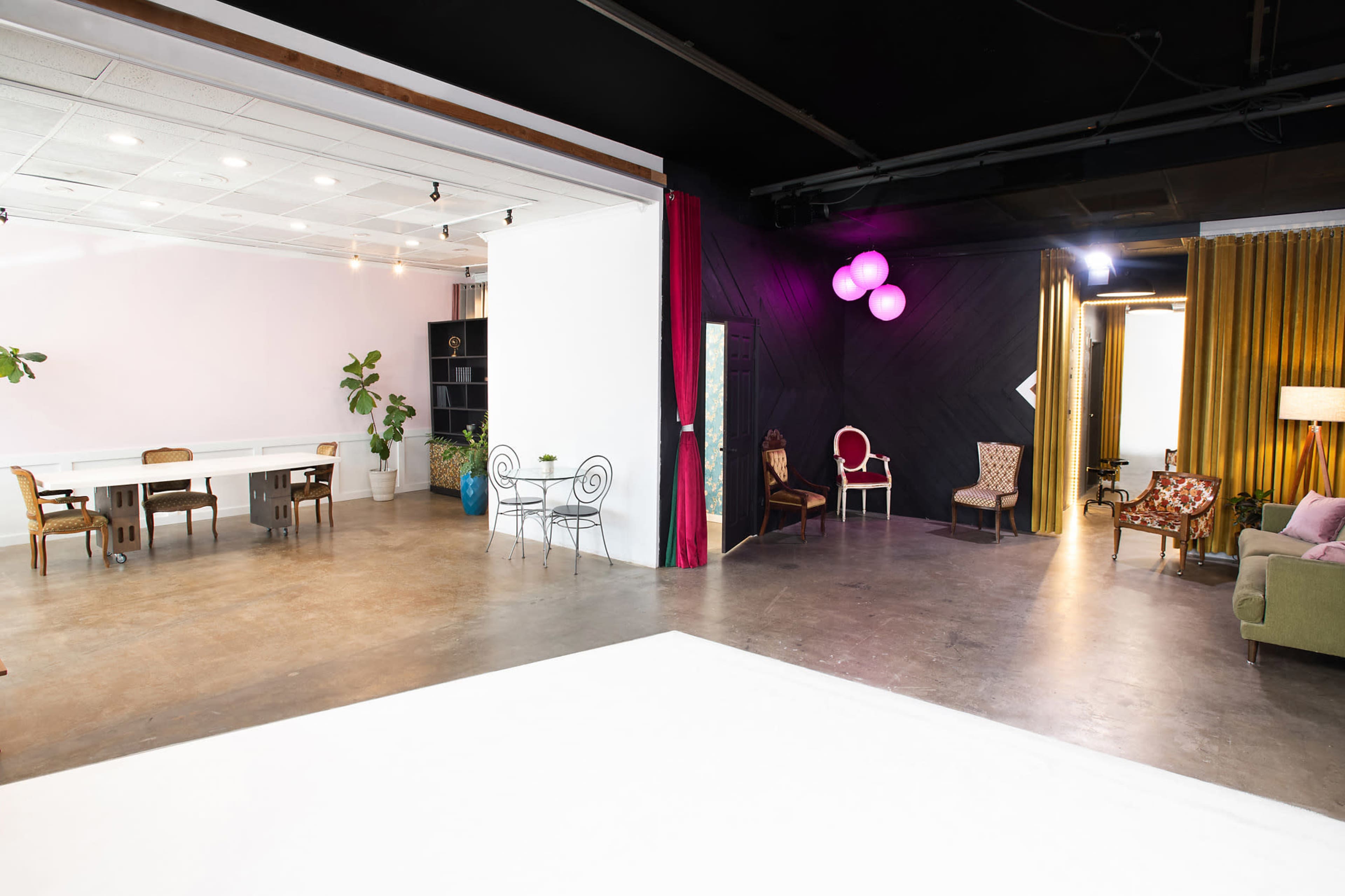 The image shows a spacious studio divided into two areas, featuring a dining setup with chairs and a table on one side and a cozy seating arrangement with plants and decorative curtains on the other.