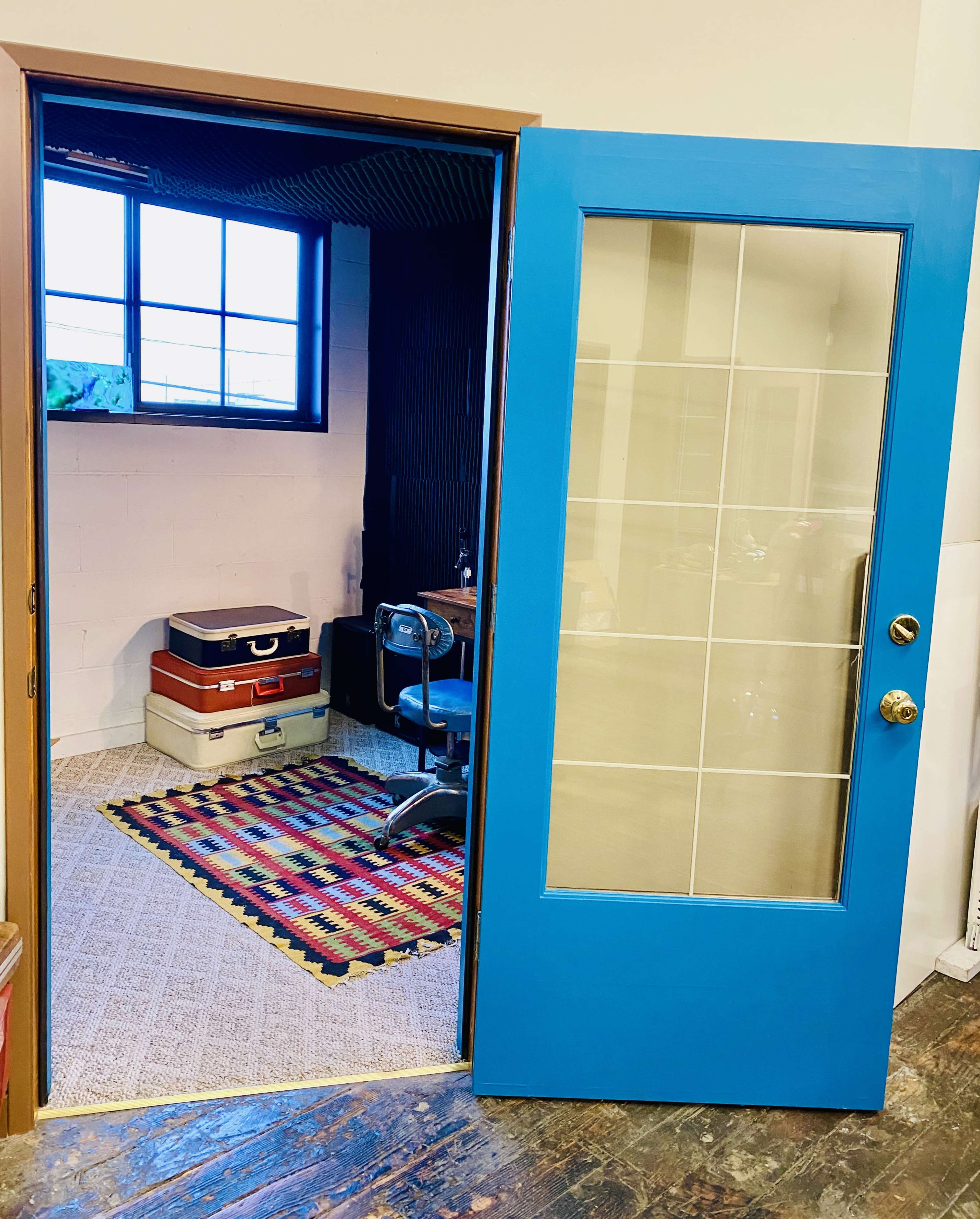 A blue door with glass panels opens to reveal a room featuring a chair, a desk, and suitcases on a patterned rug.