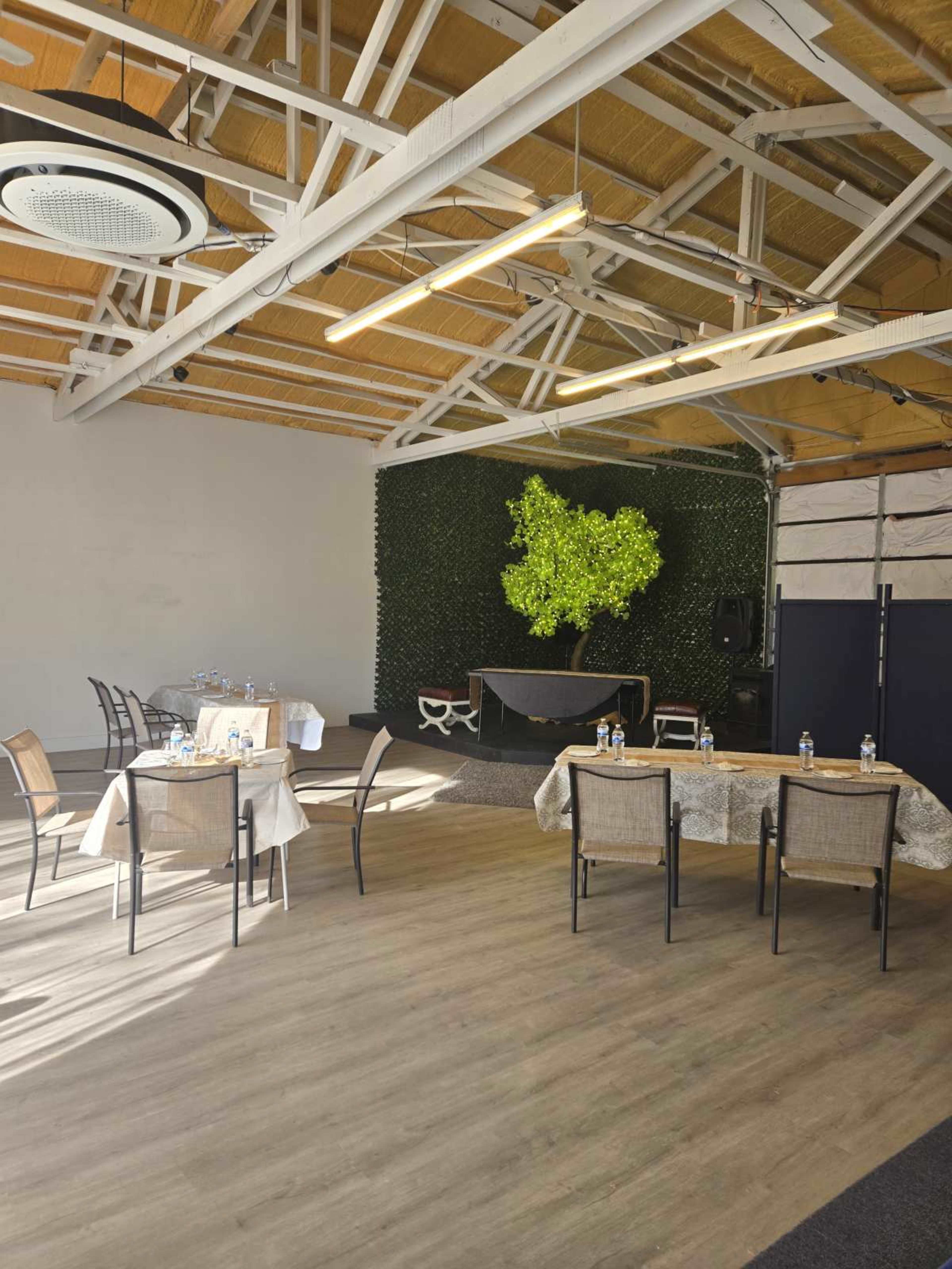 The image shows a spacious indoor area with tables and chairs arranged, featuring a green wall and a stage setup at one end.