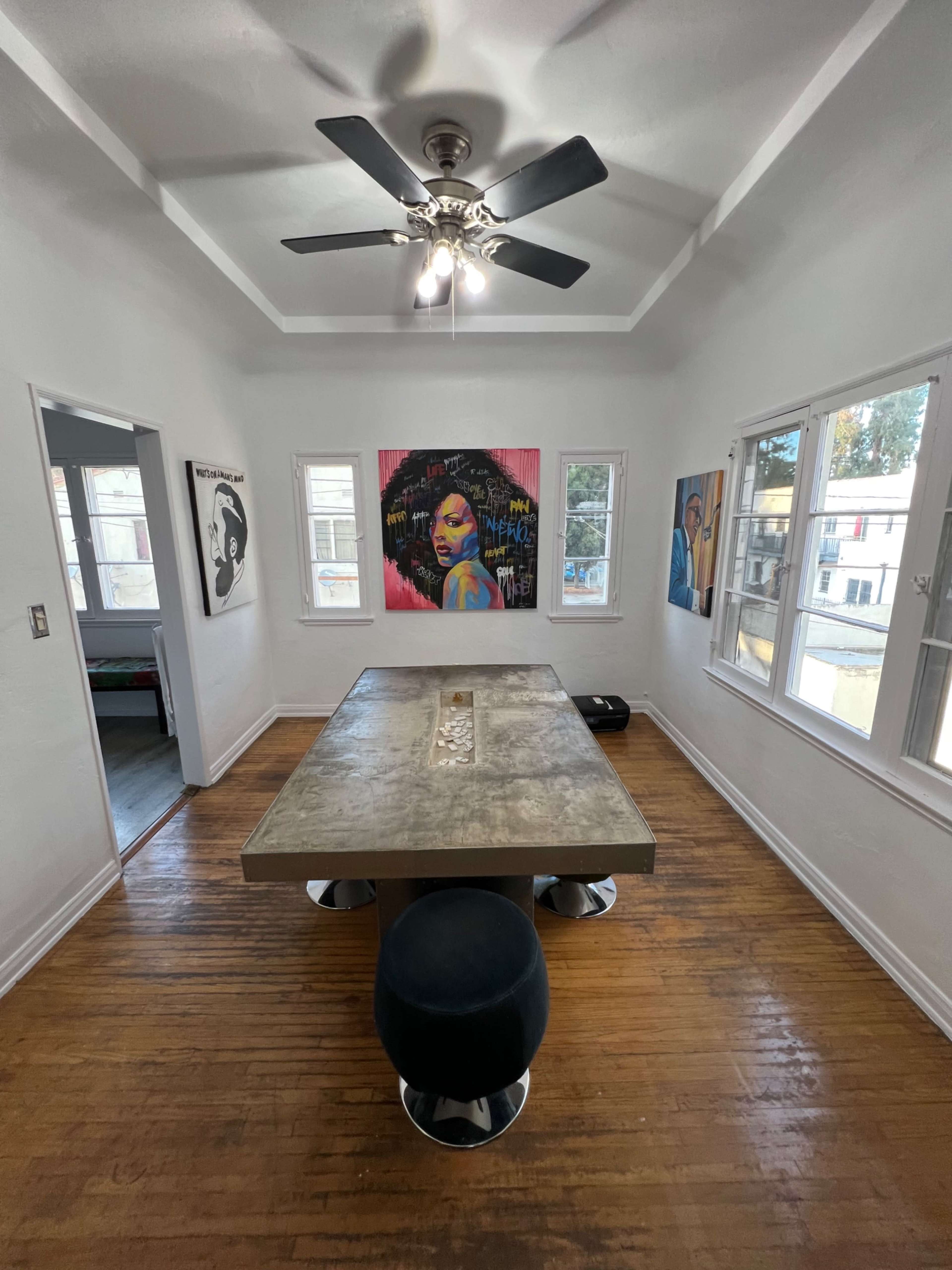 A sunlight-filled room features a large concrete table with a central centerpiece, surrounded by windows and adorned with colorful artwork on the walls.