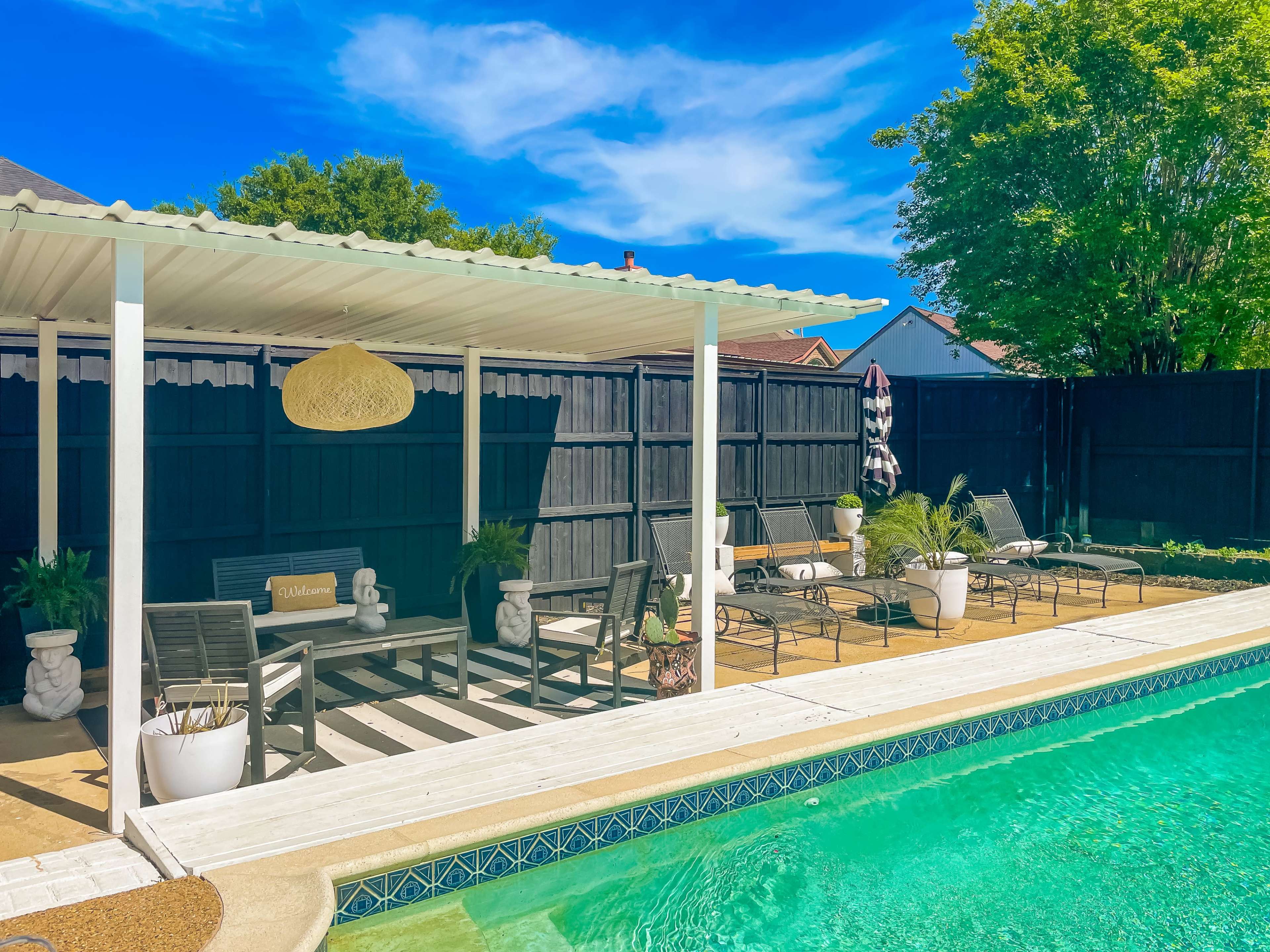 A swimming pool area with a covered seating space, lounge chairs, potted plants, and a clear blue sky.