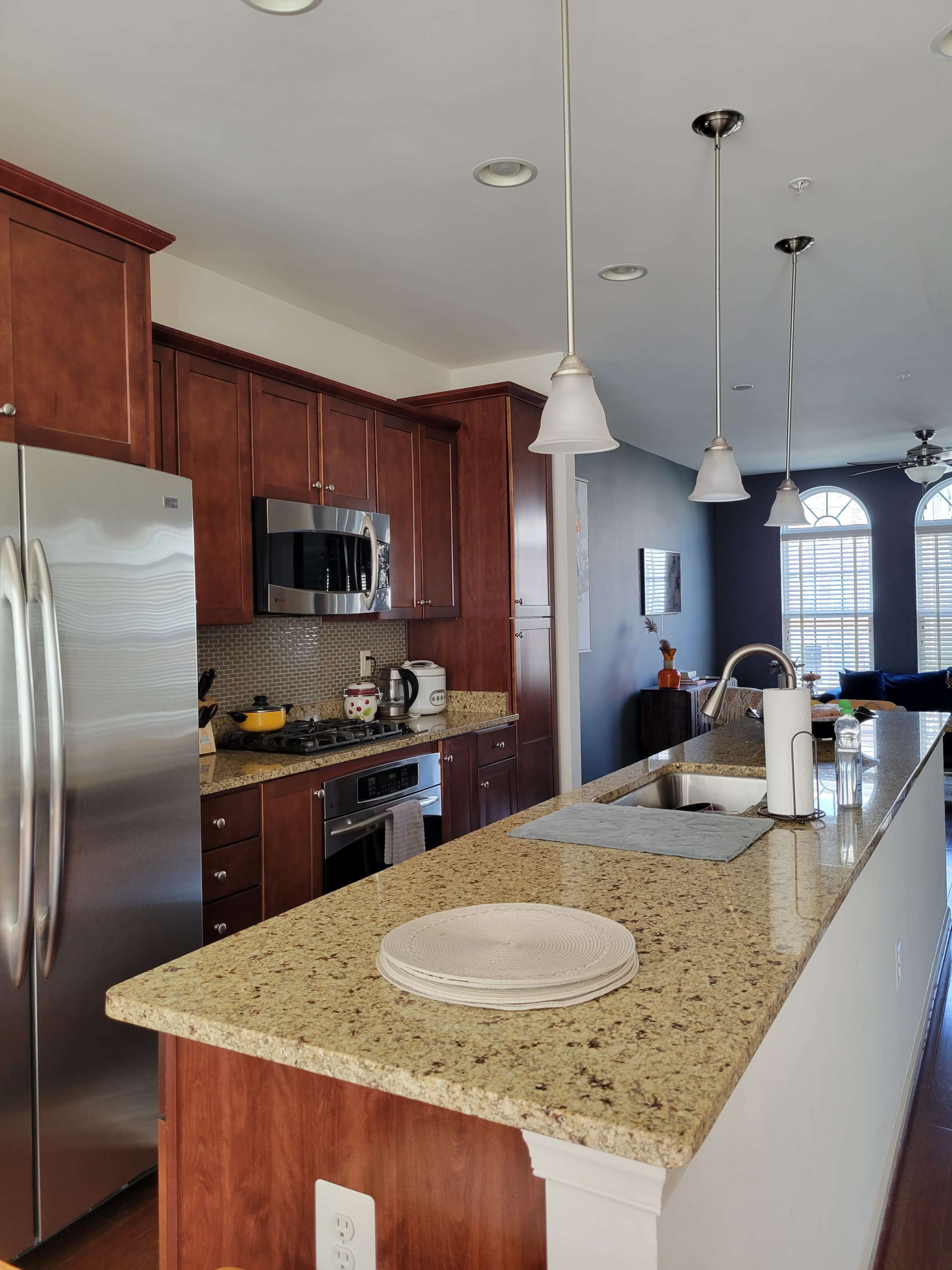A modern kitchen with wooden cabinetry, stainless steel appliances, and a granite countertop featuring a small sink and stacked dishes.
