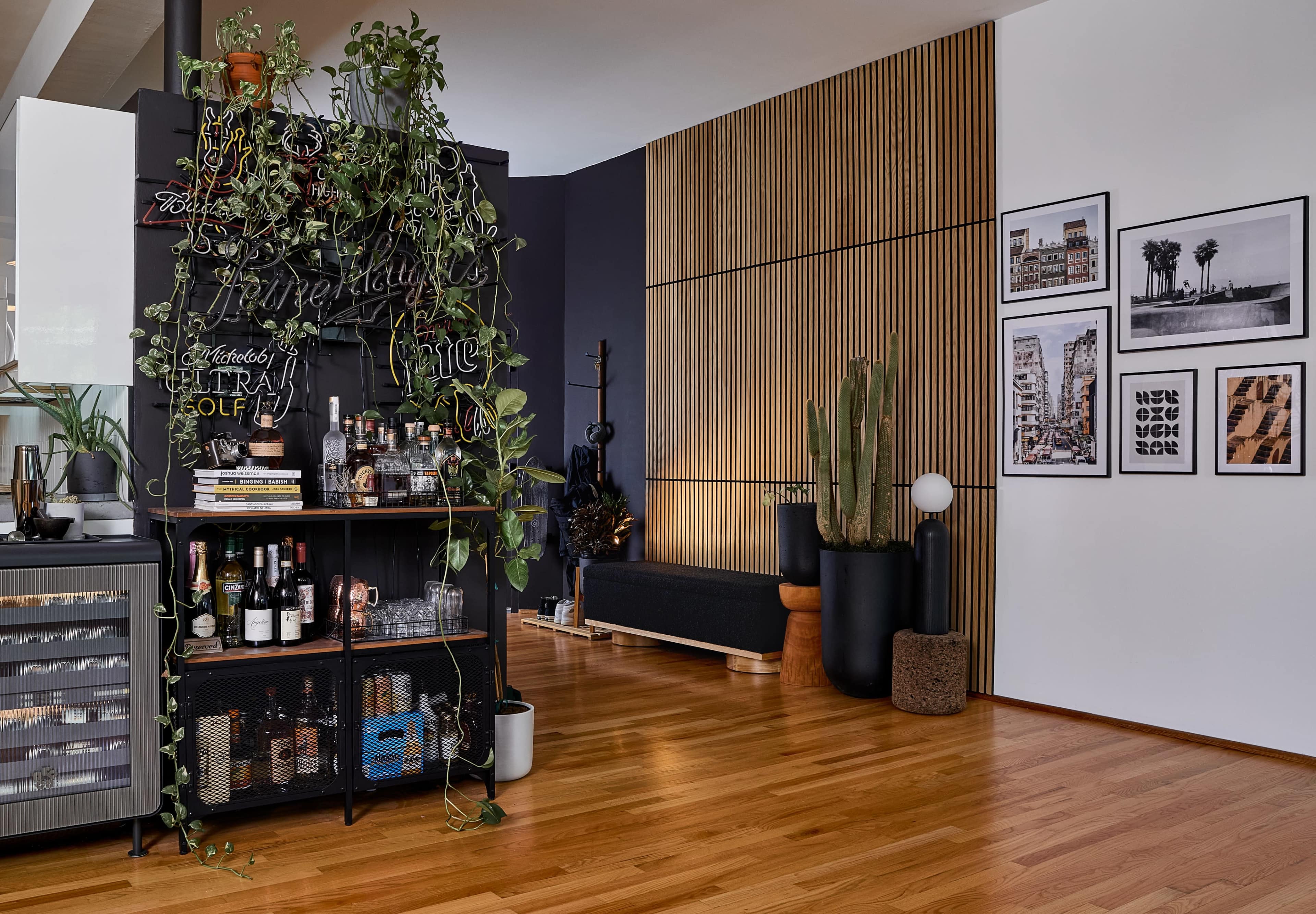 The image depicts a modern interior space featuring a bar cart surrounded by plants, wooden flooring, and a wall adorned with framed photographs.