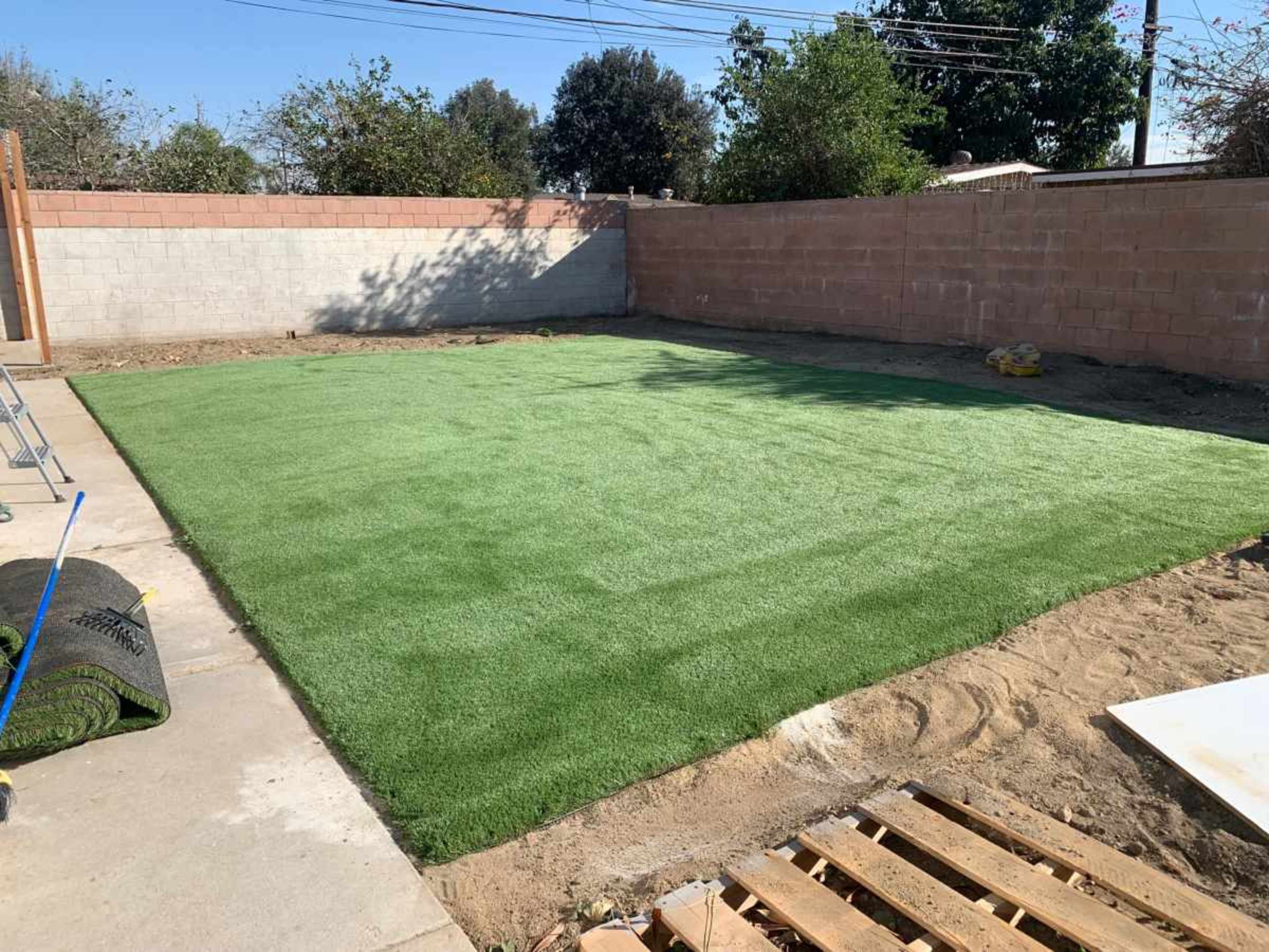 A freshly installed patch of artificial grass in a backyard, surrounded by a brick wall and sparse vegetation.