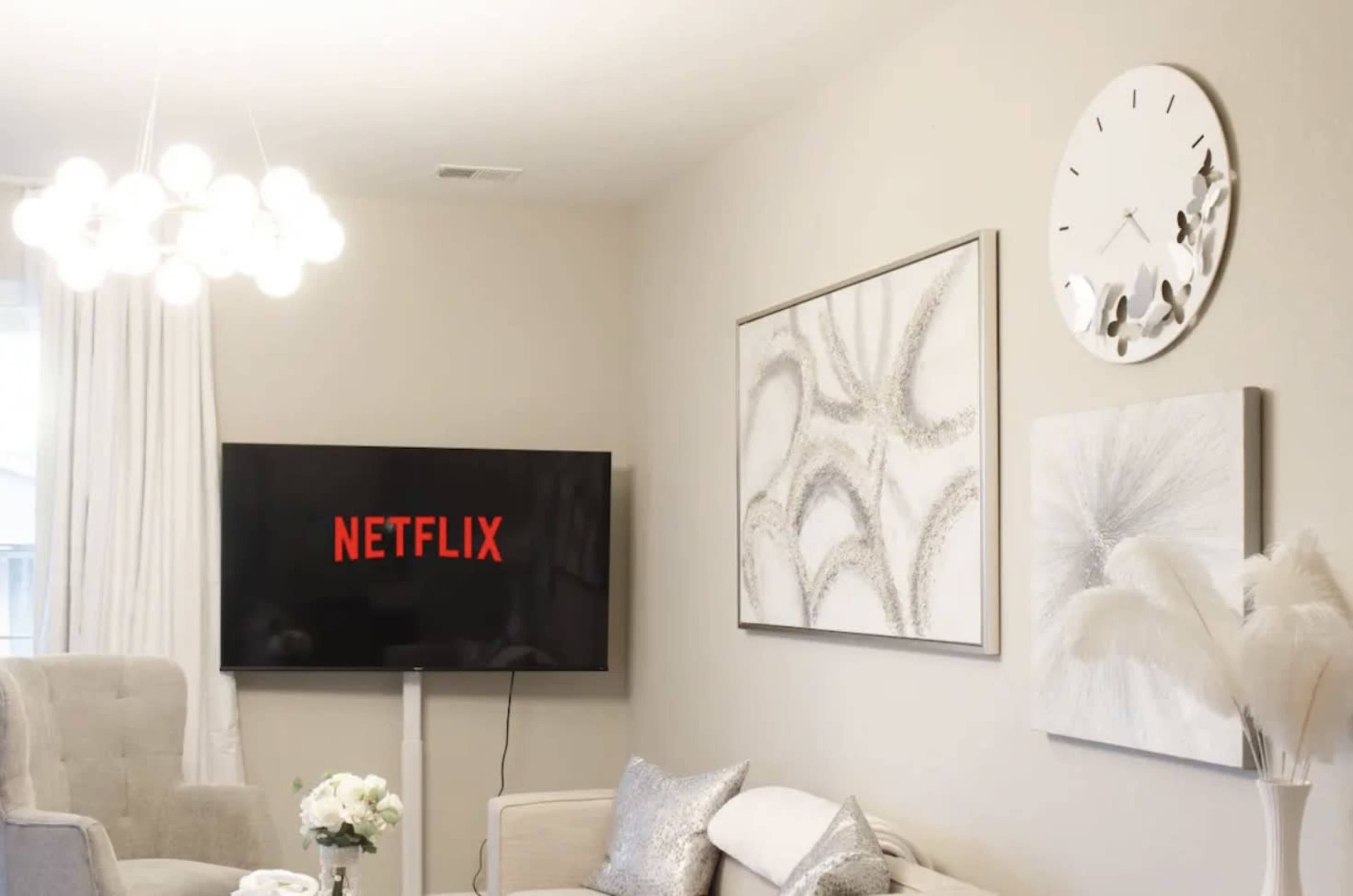 A living room features a television displaying the Netflix logo, with modern artwork and a clock on the walls, and a light fixture hanging above.