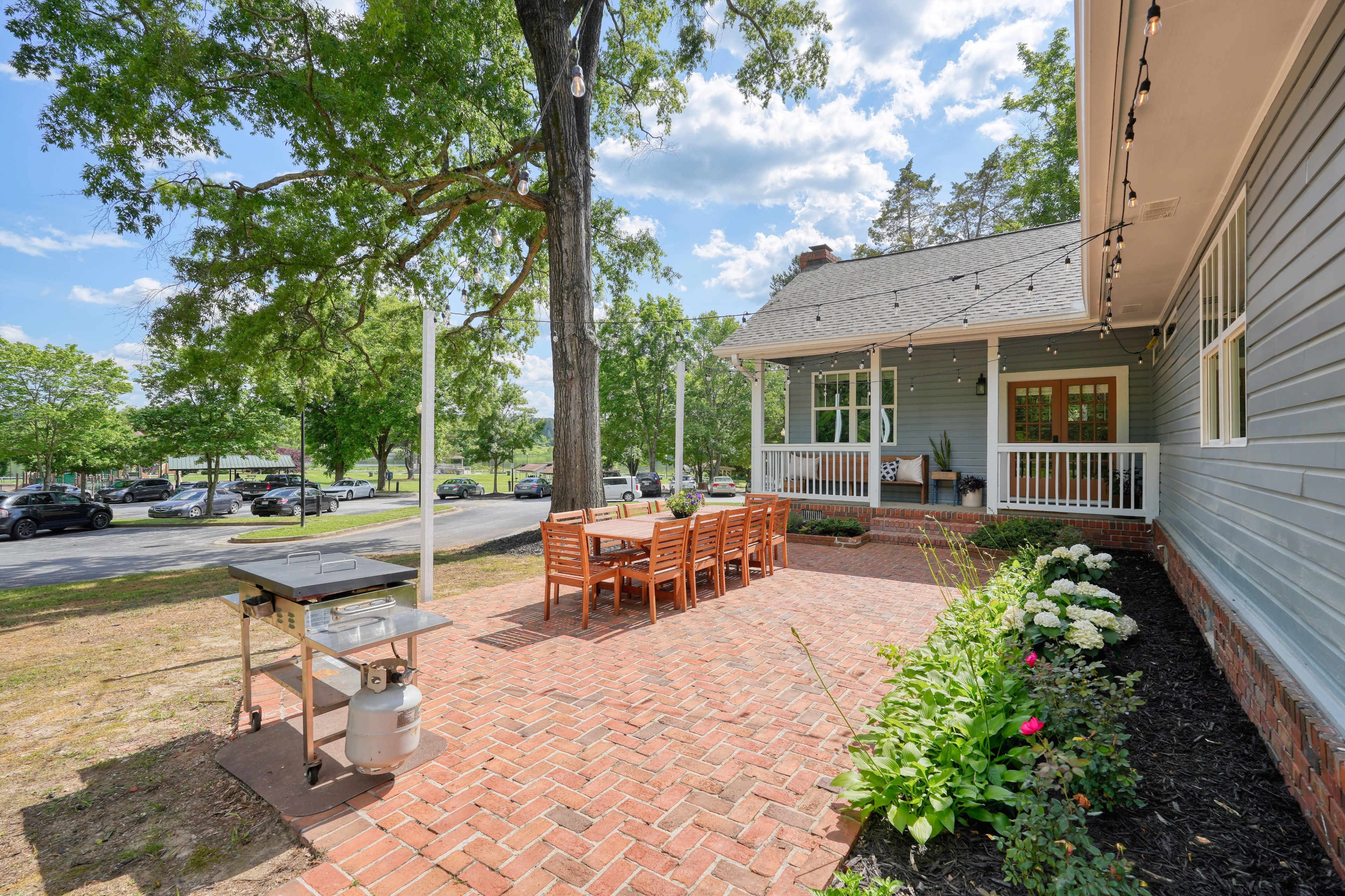 A brick patio with outdoor dining furniture and a grill is situated beside a house, surrounded by green trees and a parking area in the background.