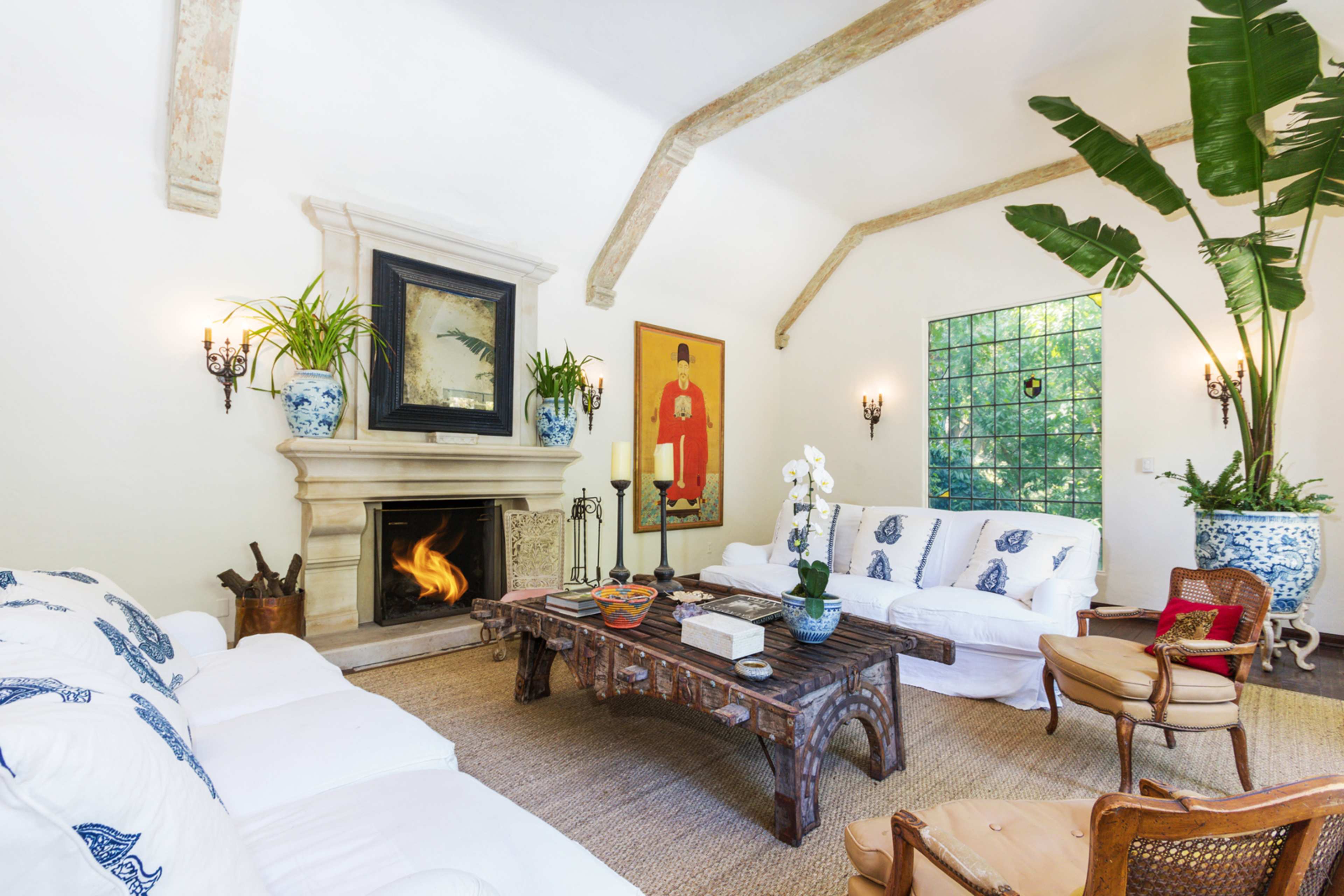 A spacious living room features a central coffee table in front of a white sofa, with a fireplace framed by artwork and potted plants decorating the space.