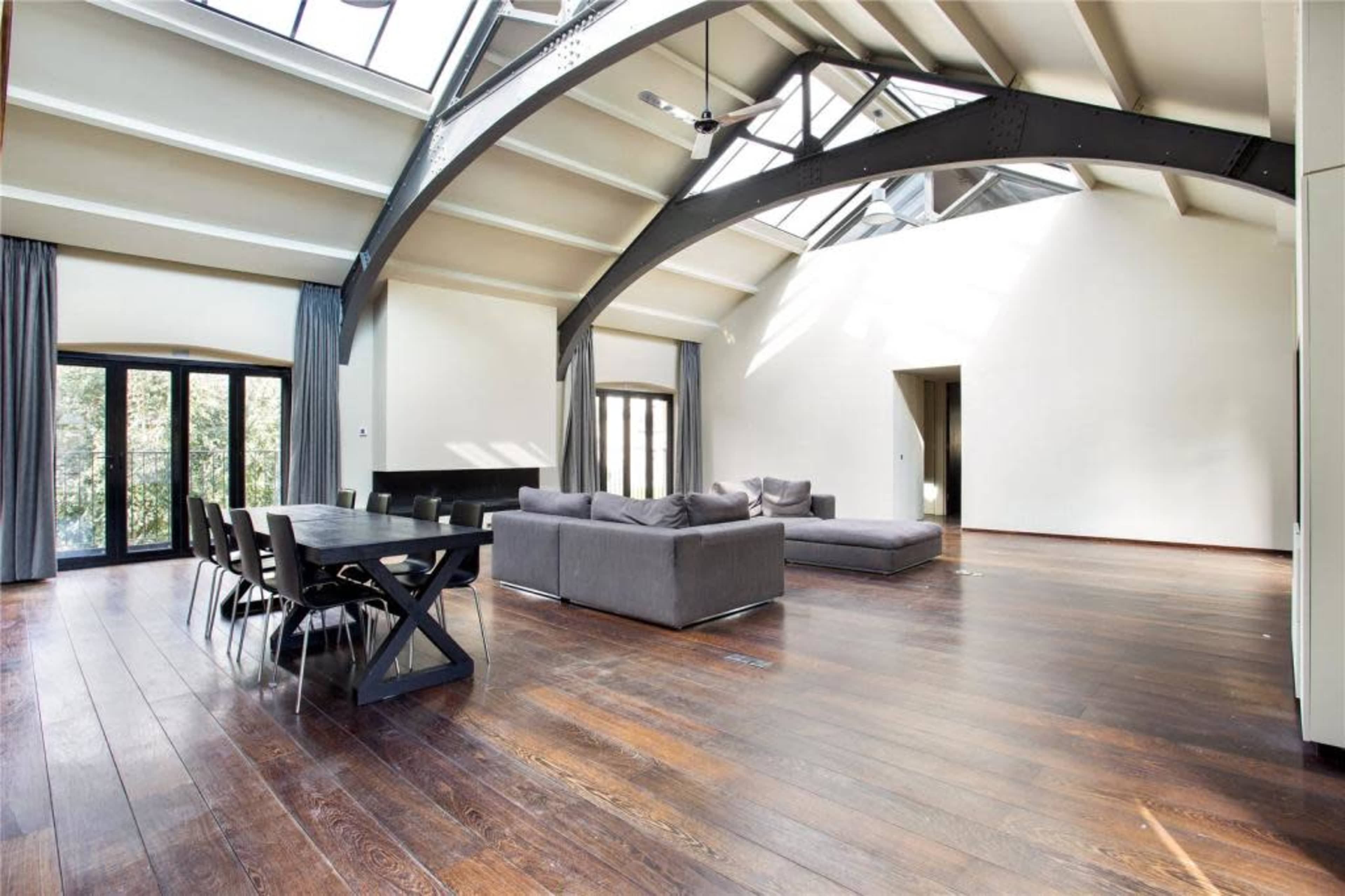 A spacious, modern living room featuring large windows, high ceilings with exposed beams, and a wooden floor, furnished with a sectional sofa and a dining table.